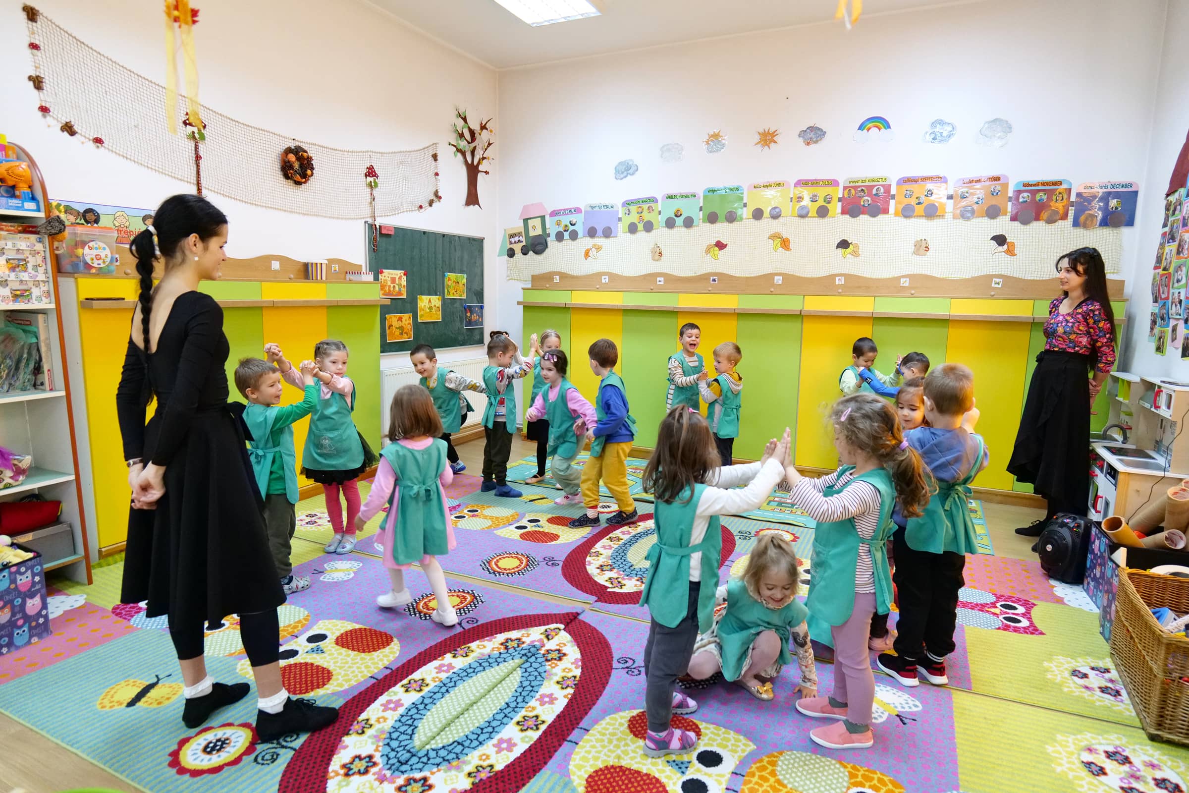 Óvodások énekelnek és táncolnak a csoportszoba színes szőnyegén, két pedagógus irányításával.