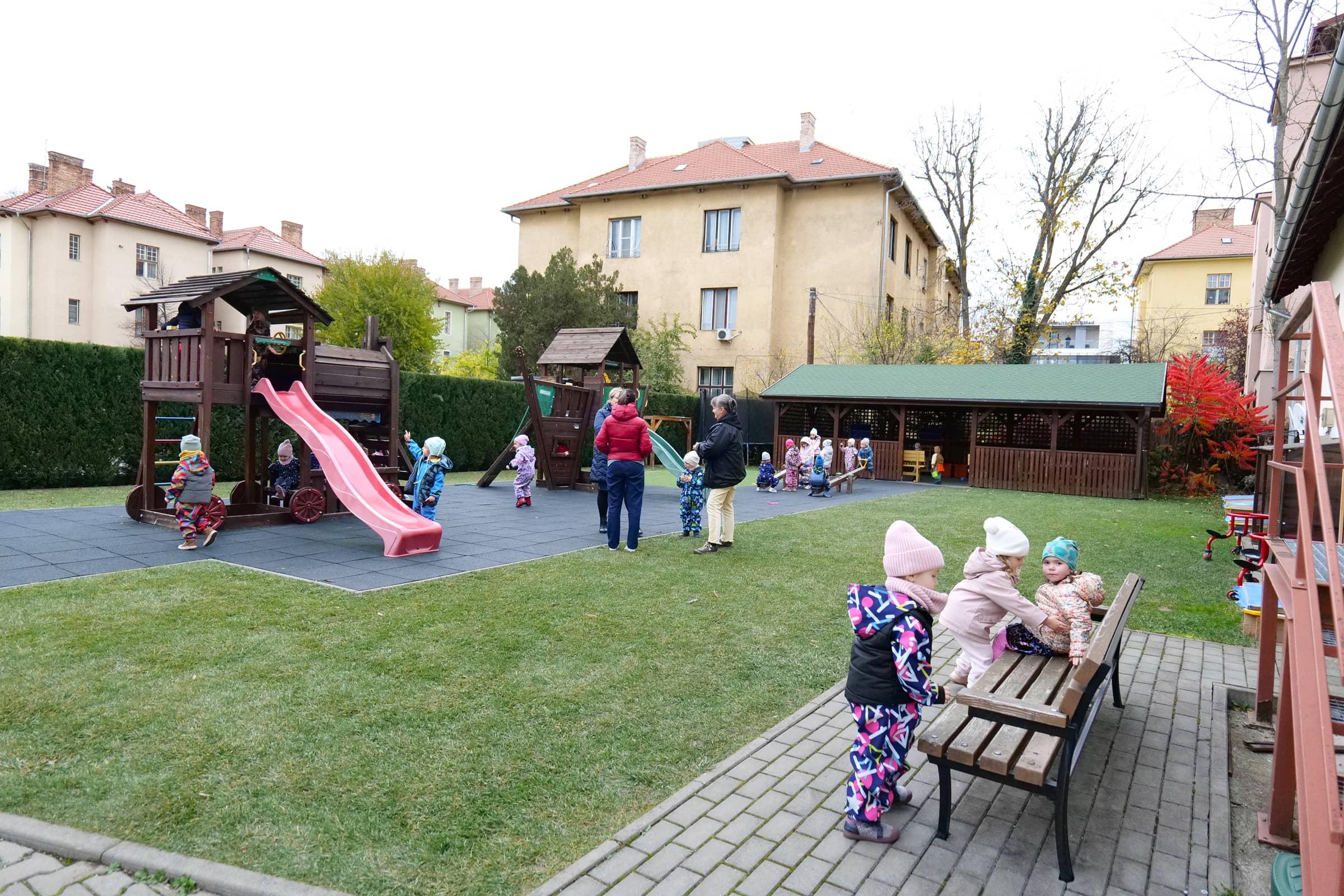 Óvodások az óvoda udvarán csúszdáznak, másznak és beszélgetnek egymással, a pedagógusok felügyeletével.