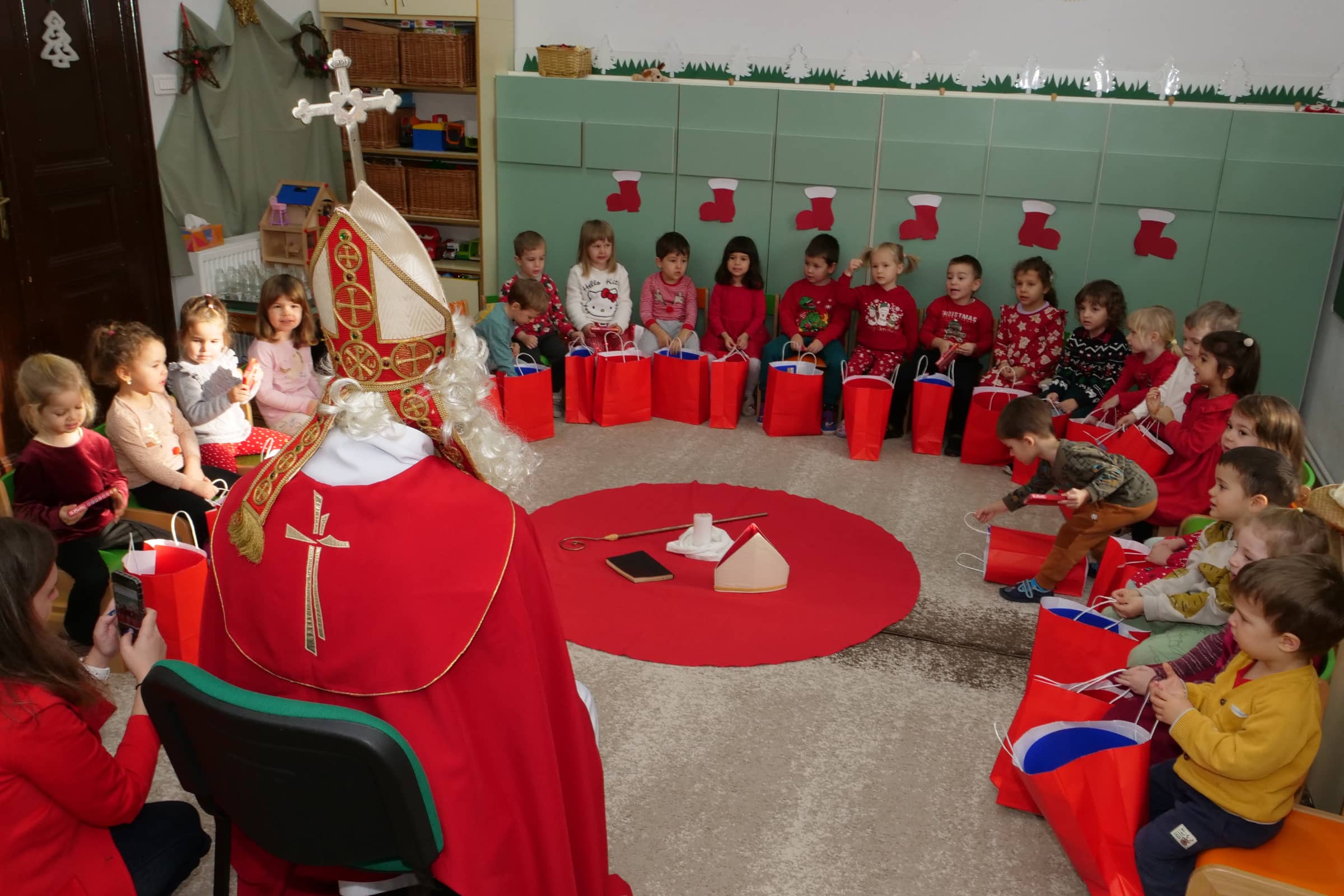 Szent Miklós látogatása az óvodában: a gyermekek körben ülve ajándékcsomagokkal hallgatják Szent Miklóst