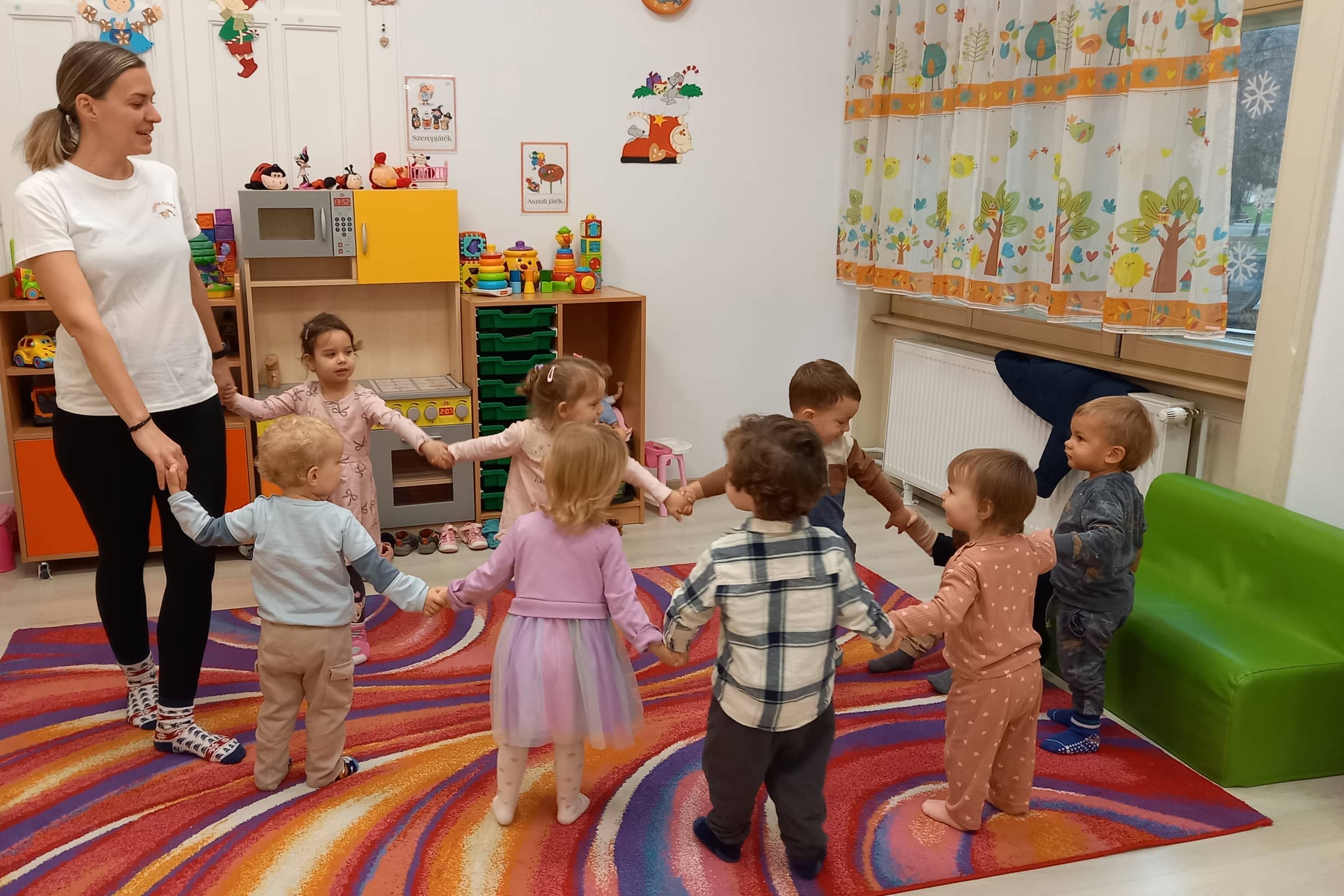 Mozgásfejlesztő foglalkozás az óvodában: a kisgyermekek körben állva, kézen fogva mozognak a pedagógus irányításával