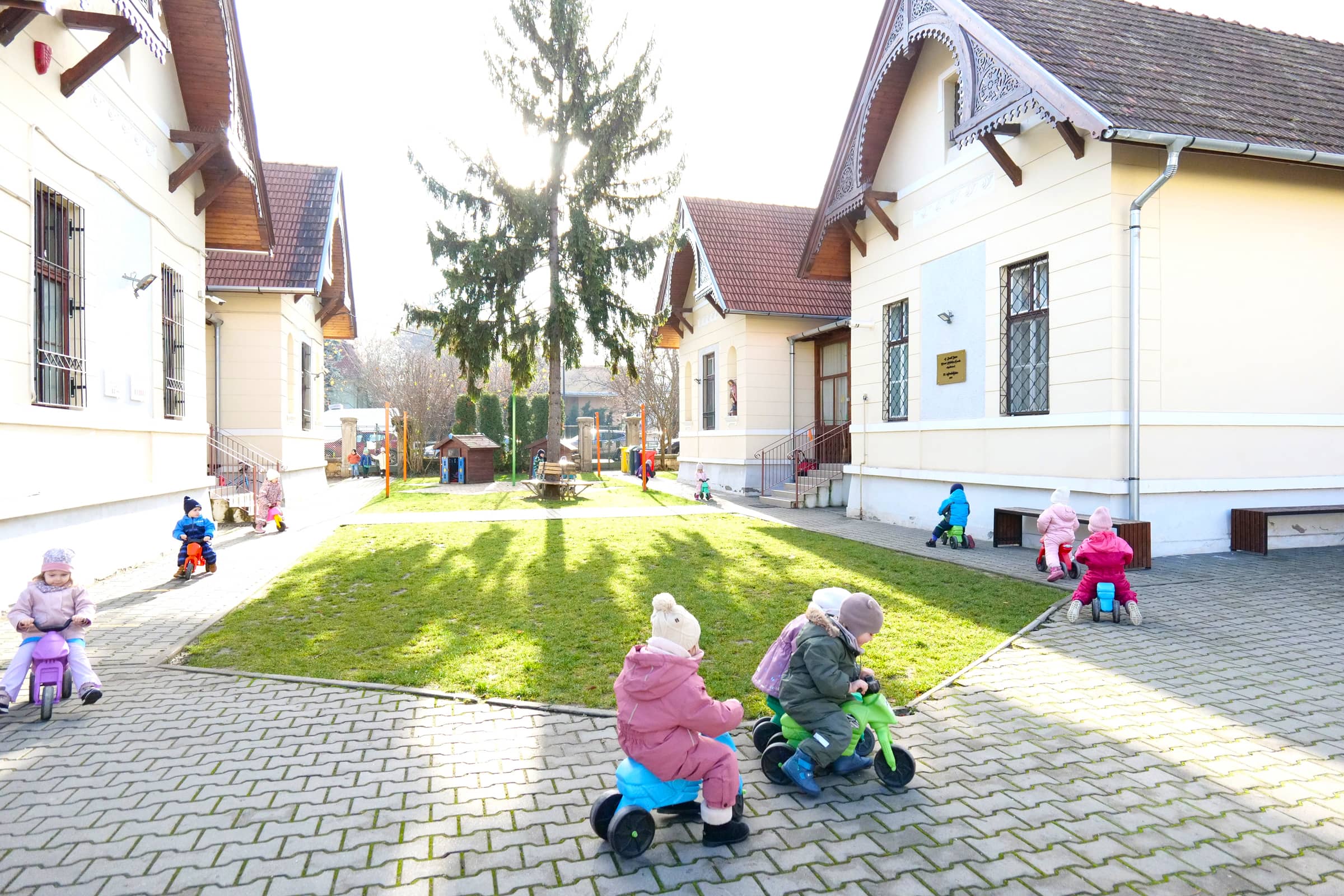 A gyerekek kismotorokkal és futóbiciklikkel játszanak az óvoda udvarán.