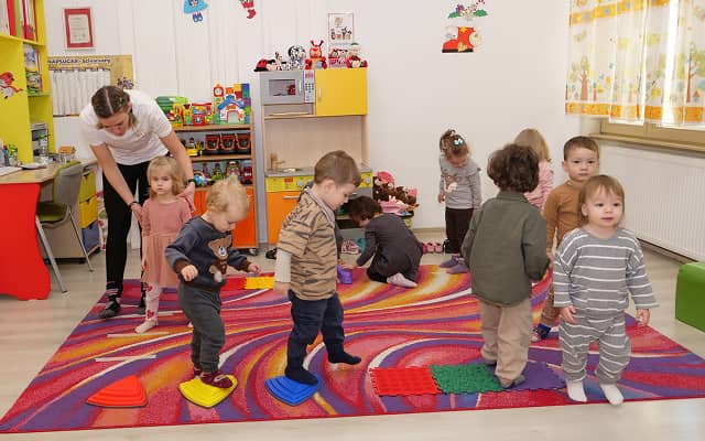 Gyermekek játékos mozgásos feladatot végeznek színes akadálypályán egy bölcsődei teremben, pedagógus felügyelete mellett.