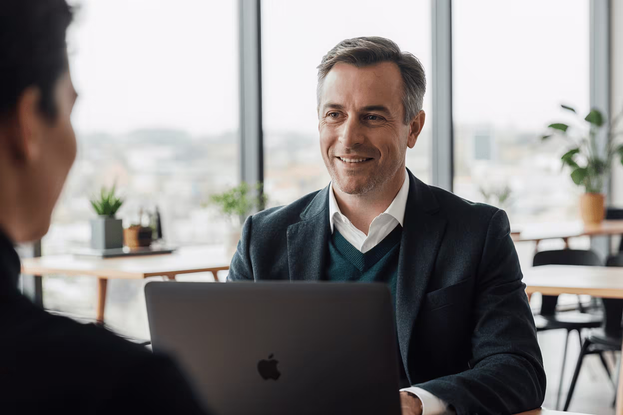 Homme professionnel de 40 ans dans un open space lumineux à Lausanne, discutant avec un collègue, MacBook Pro devant lui.