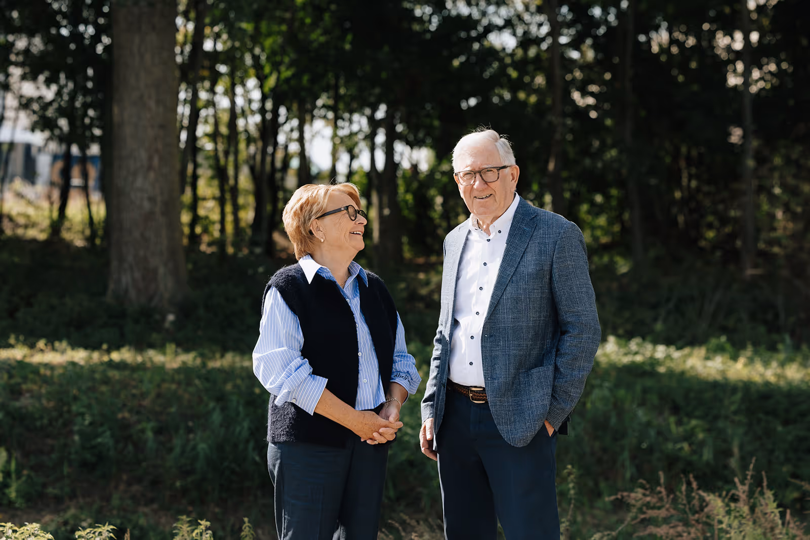 Twee oudere mensen staan buiten in een zonovergoten bosrijke omgeving, glimlachend en pratend.