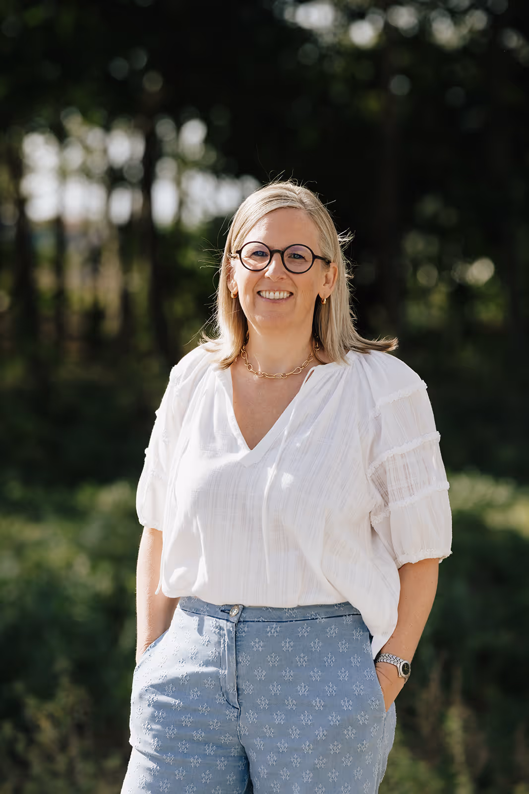 Vrouw met blond haar en bril, gekleed in een witte blouse en lichtblauwe broek met patroon, glimlacht buiten met bomen op de achtergrond.