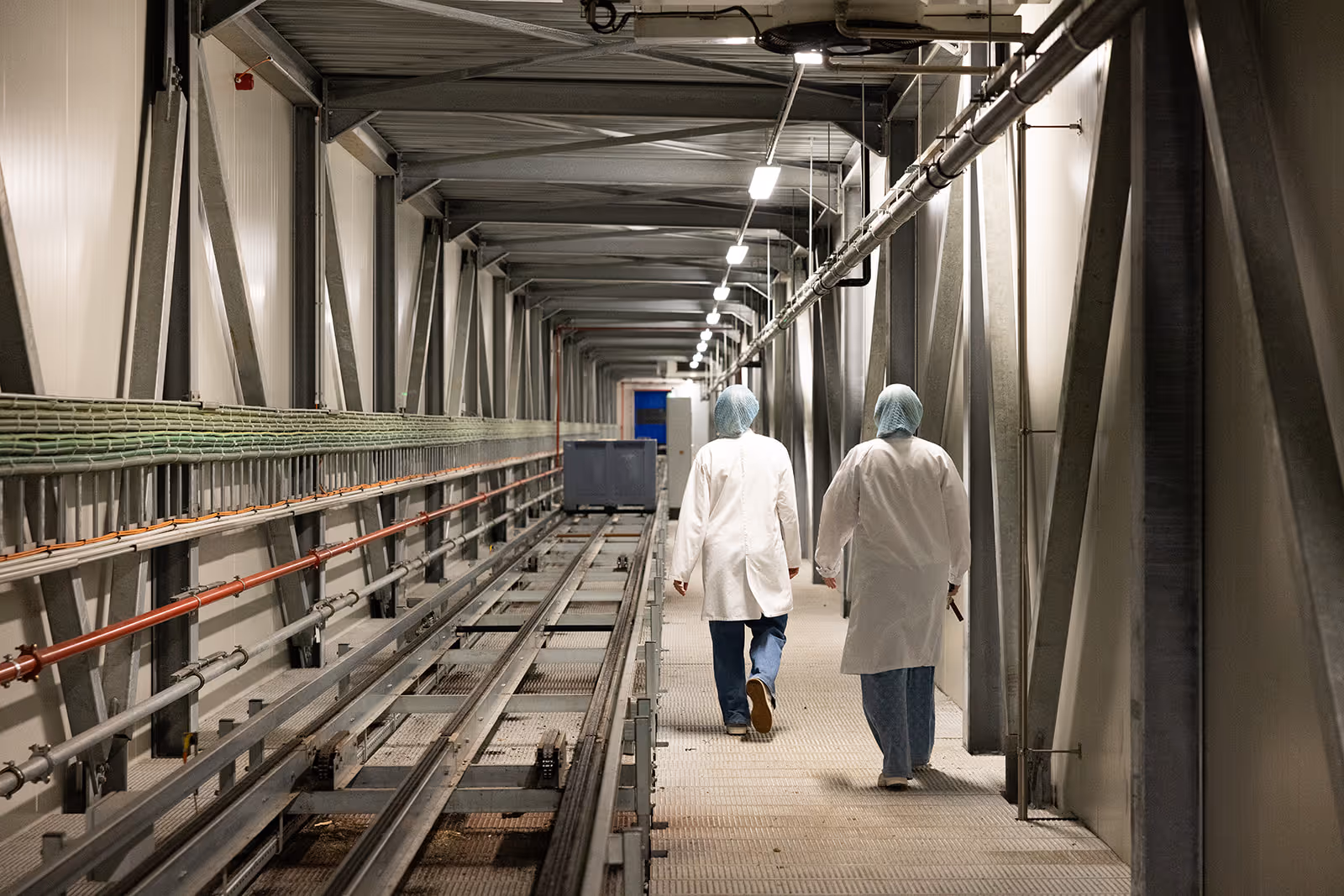 Twee mensen in witte jassen en haarnetjes lopen langs een industriële gang met metalen leidingen en rails.