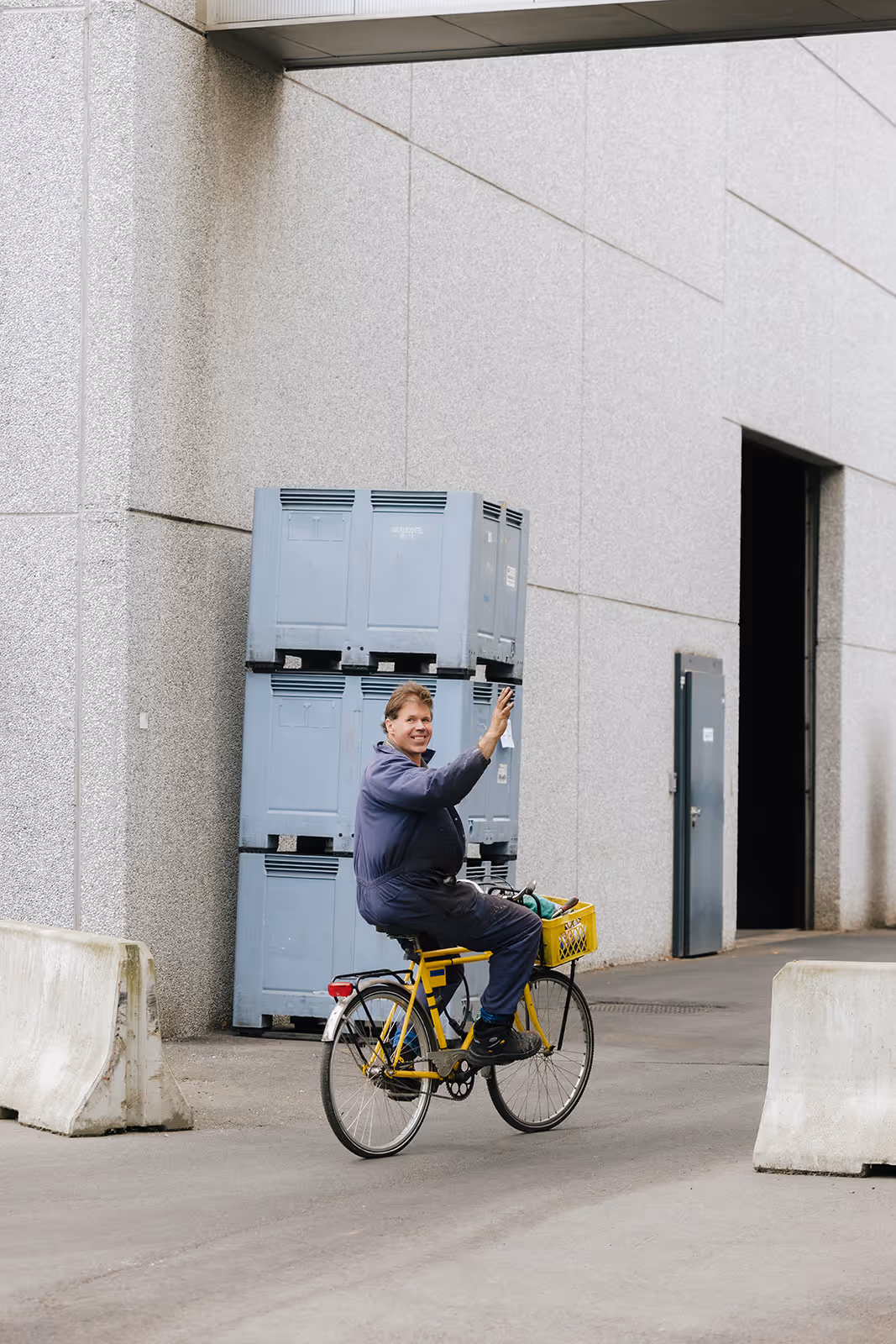 Man in work overalls waving while riding a yellow bicycle near stacked blue crates outside a building.