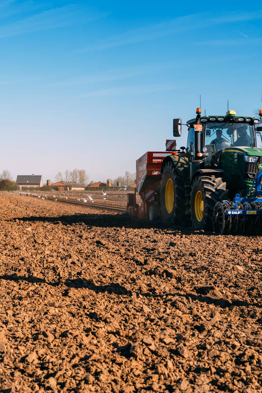 Een groene tractor werkt op een omgeploegde akker met boerderijgebouwen op de achtergrond onder een blauwe lucht.