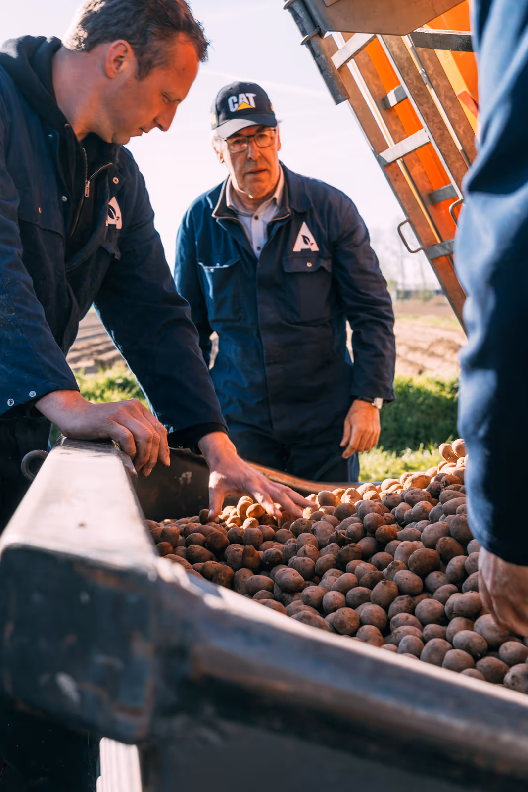 Twee mannen in blauwe werkkleding inspecteren aardappelen in een landbouwmachine buiten.