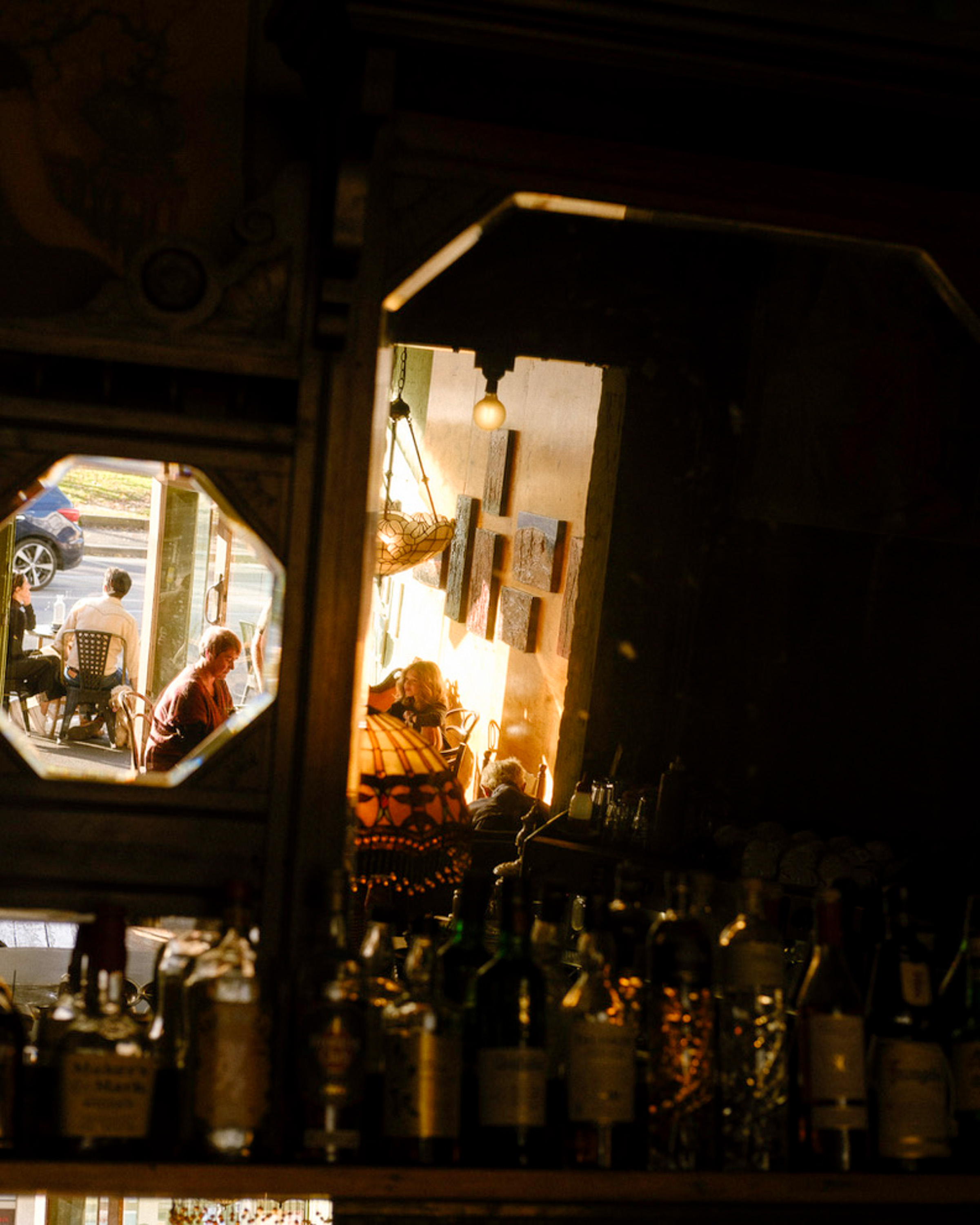Reflection of people sitting and talking inside a warmly lit café with bottles lined up on a shelf in the foreground.