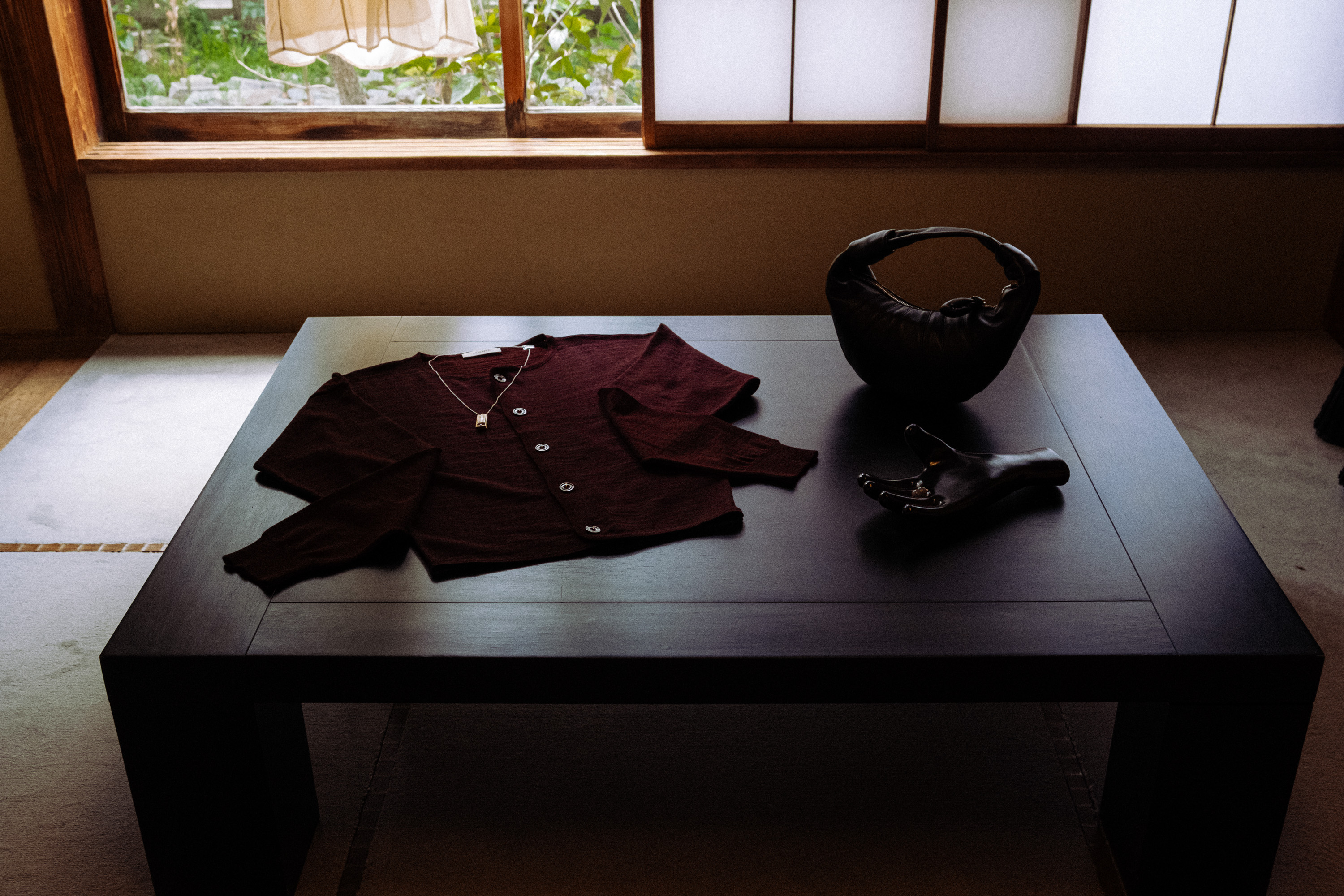Dark wooden table with a folded maroon cardigan with a necklace and two black decorative objects, set in a room with a window and beige walls.