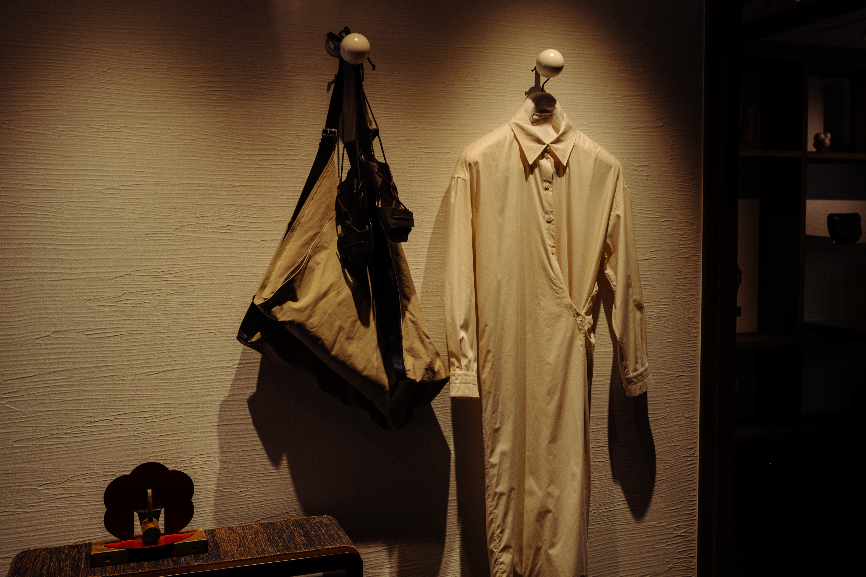 Beige long-sleeve dress and a large beige bag hanging on white wall hooks in dimly lit room.