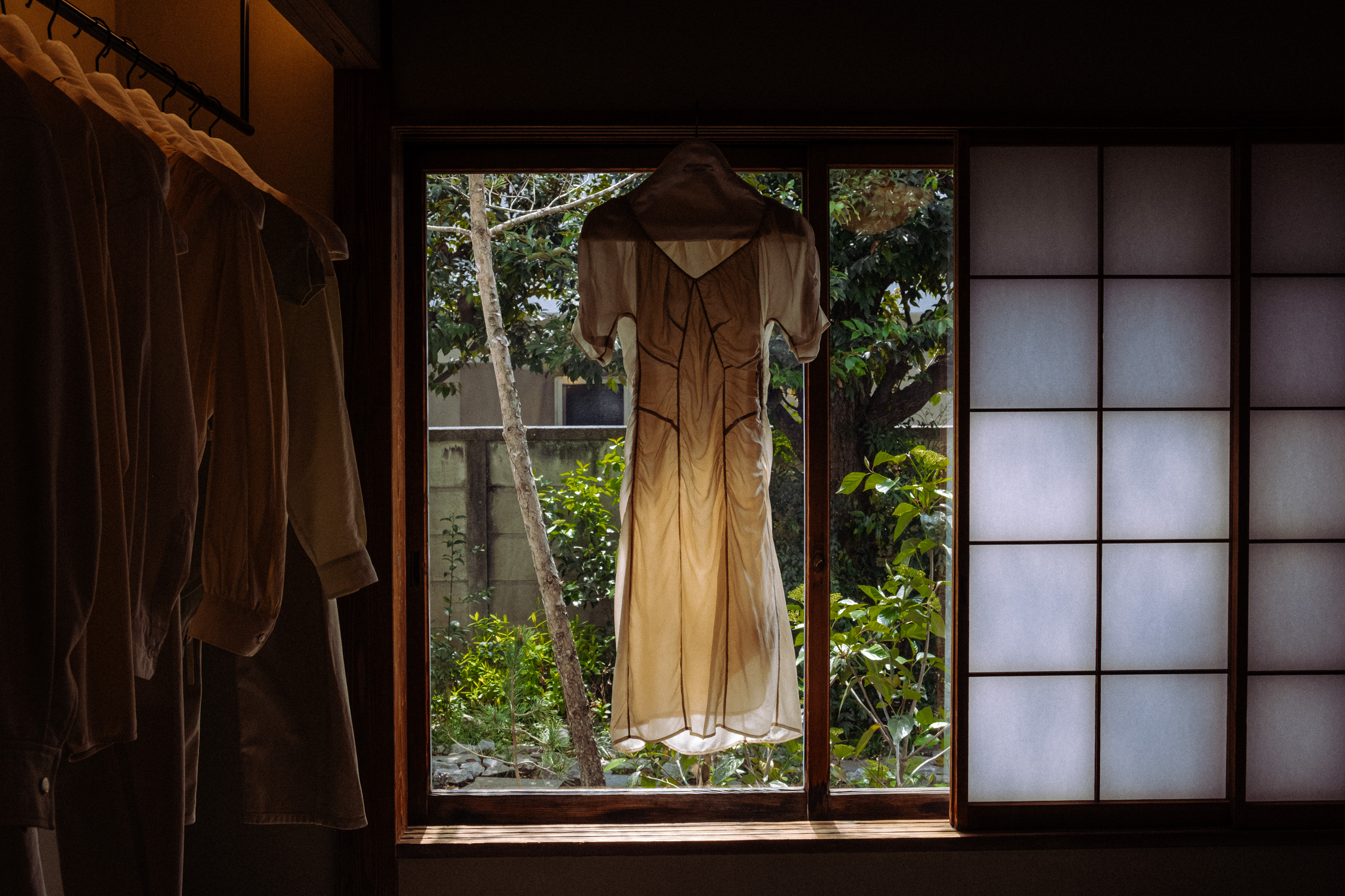 Light beige dress hanging on a window frame with green garden visible outside and soft light illuminating the dress.
