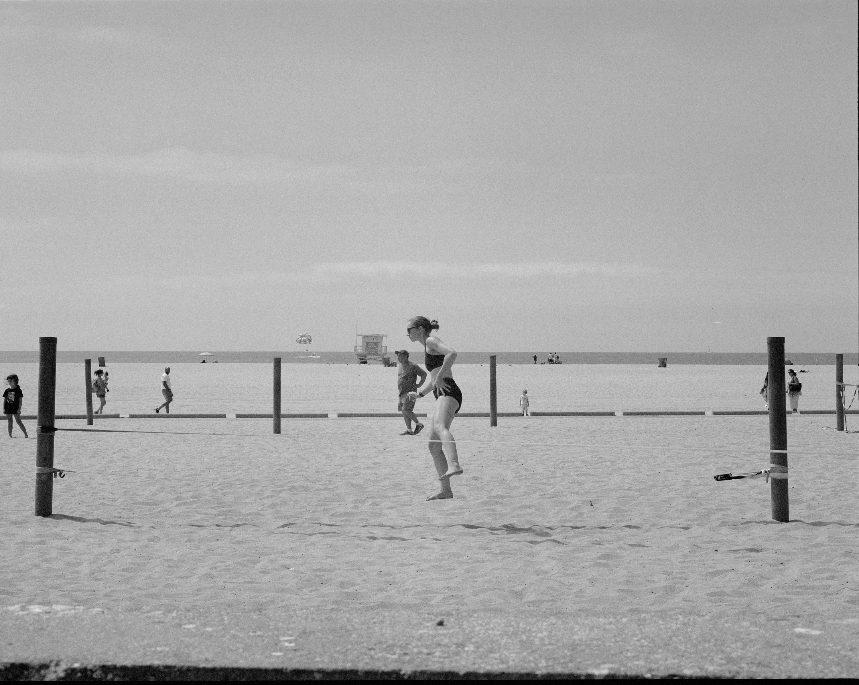 Woman in a swimsuit walking barefoot on beach sand between wooden poles, with ocean and people in the background.