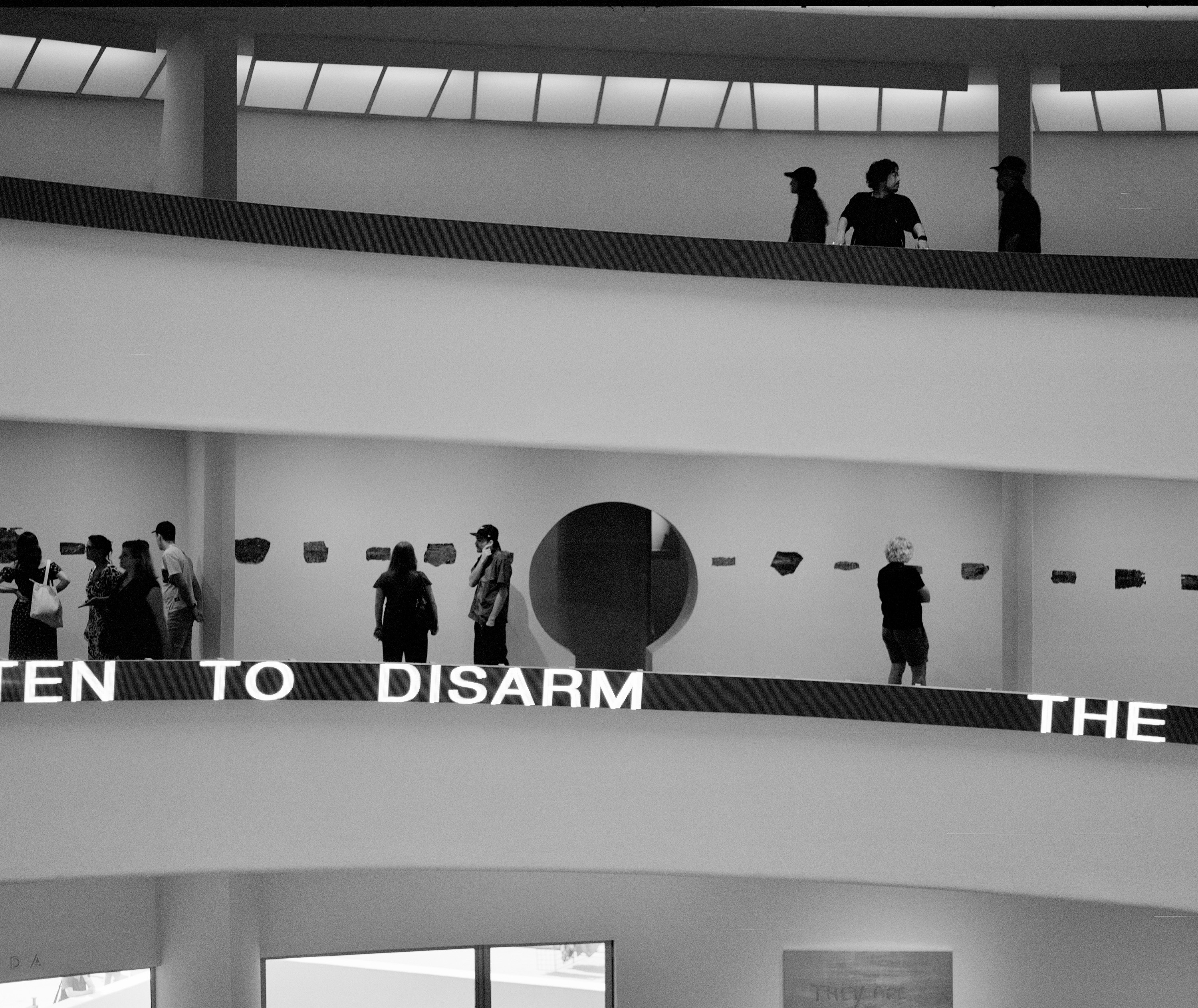 Black and white photo of people walking and standing on curved walkways inside a modern building with a partial illuminated sign displaying the words 'TO DISARM THE'.