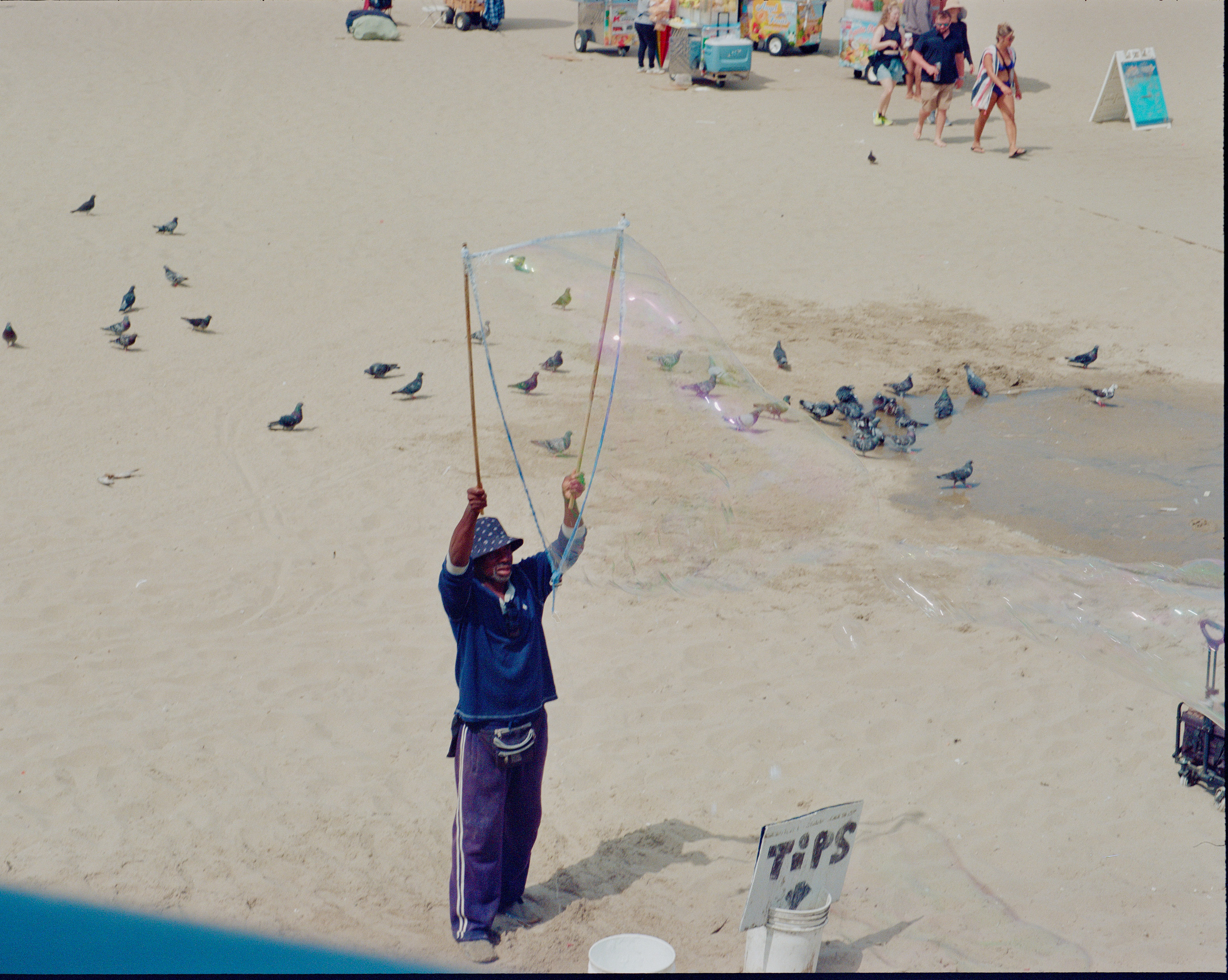 Man on sandy beach creating a large bubble using two sticks and a rope, with a tip bucket nearby and pigeons scattered around.