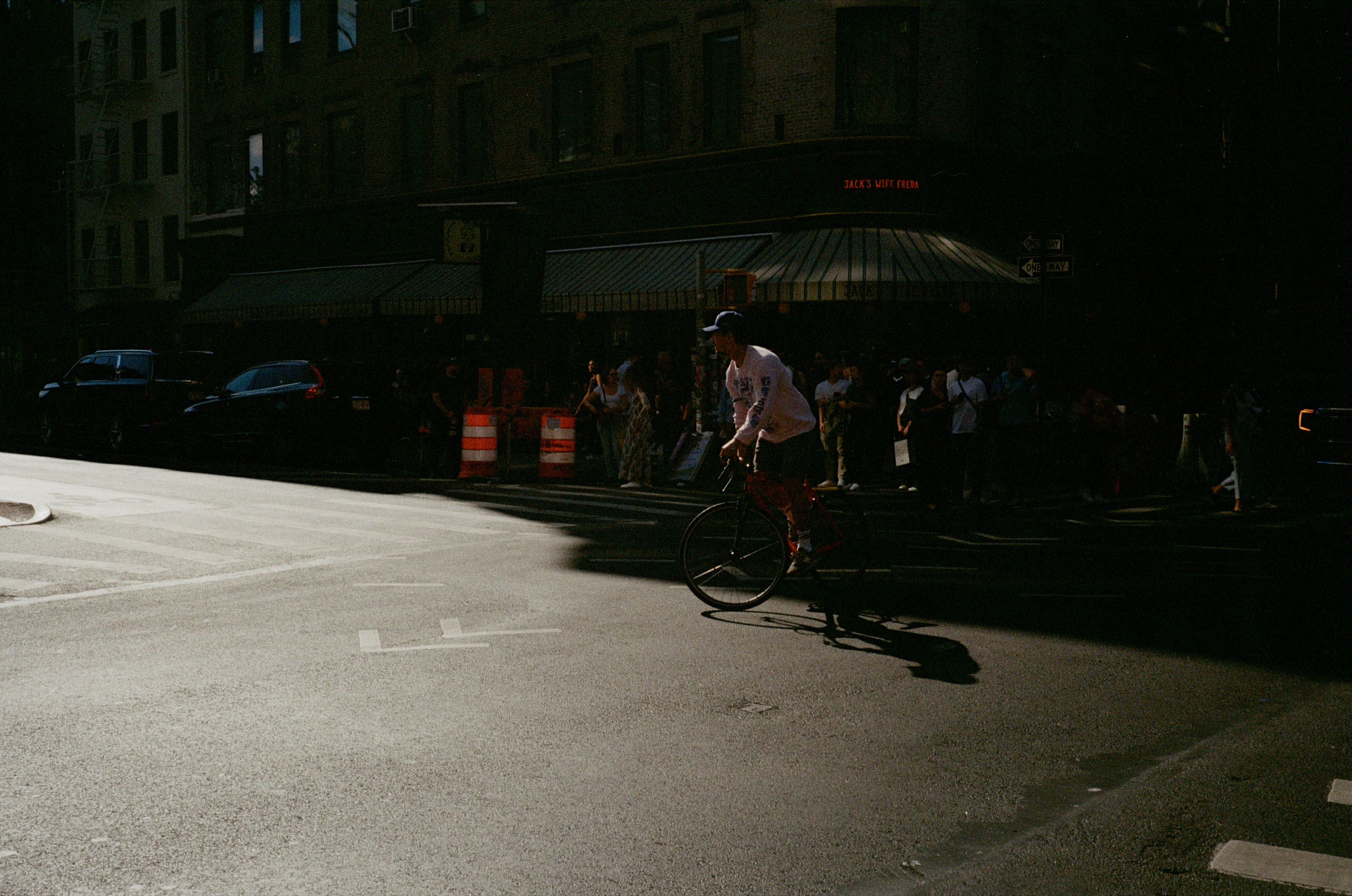 Person riding a bicycle across a sunlit city street corner with a crowd and parked cars in the shaded background.