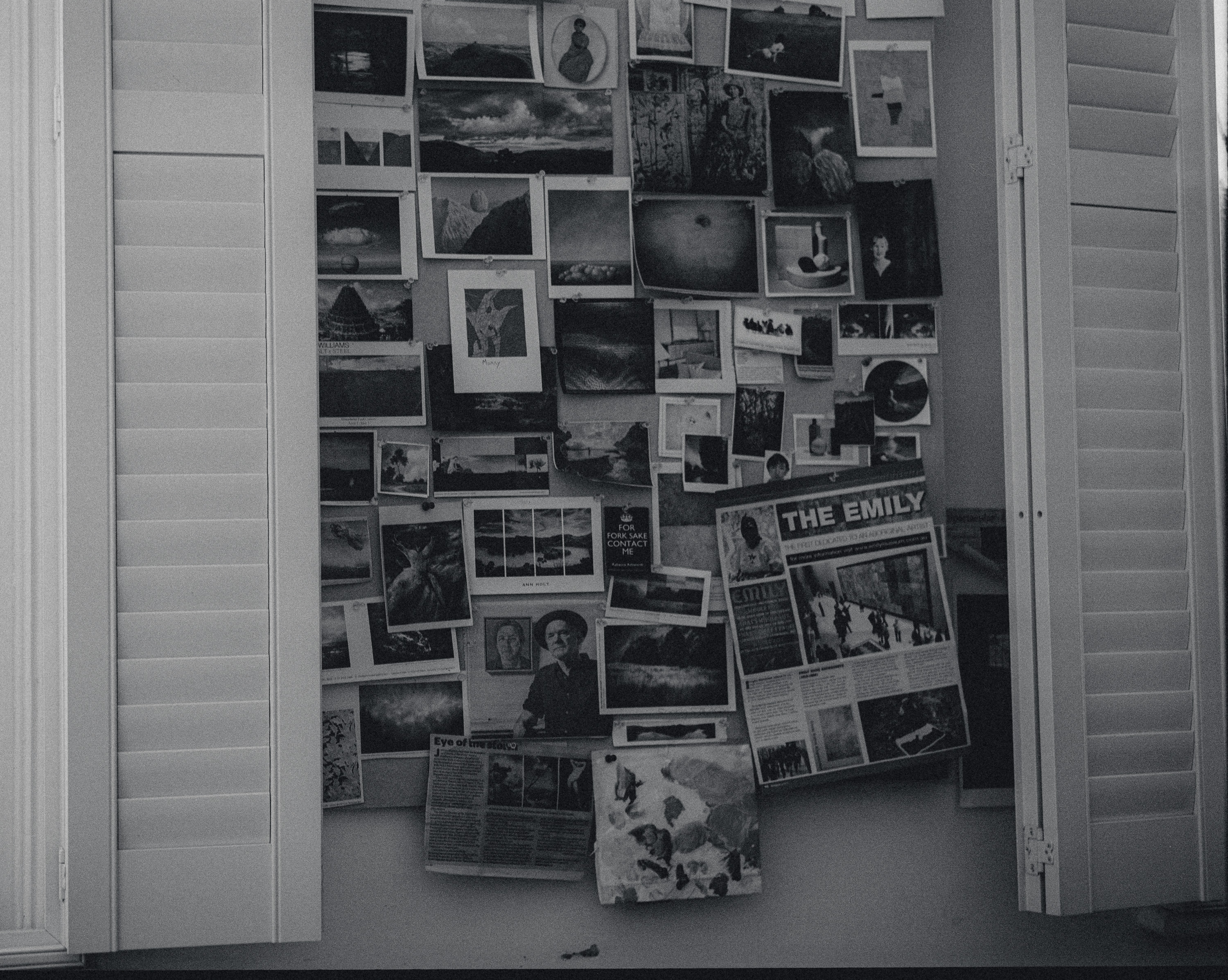 Black and white photo of a bulletin board filled with various pinned photos, clippings, and artwork, framed by open window shutters.