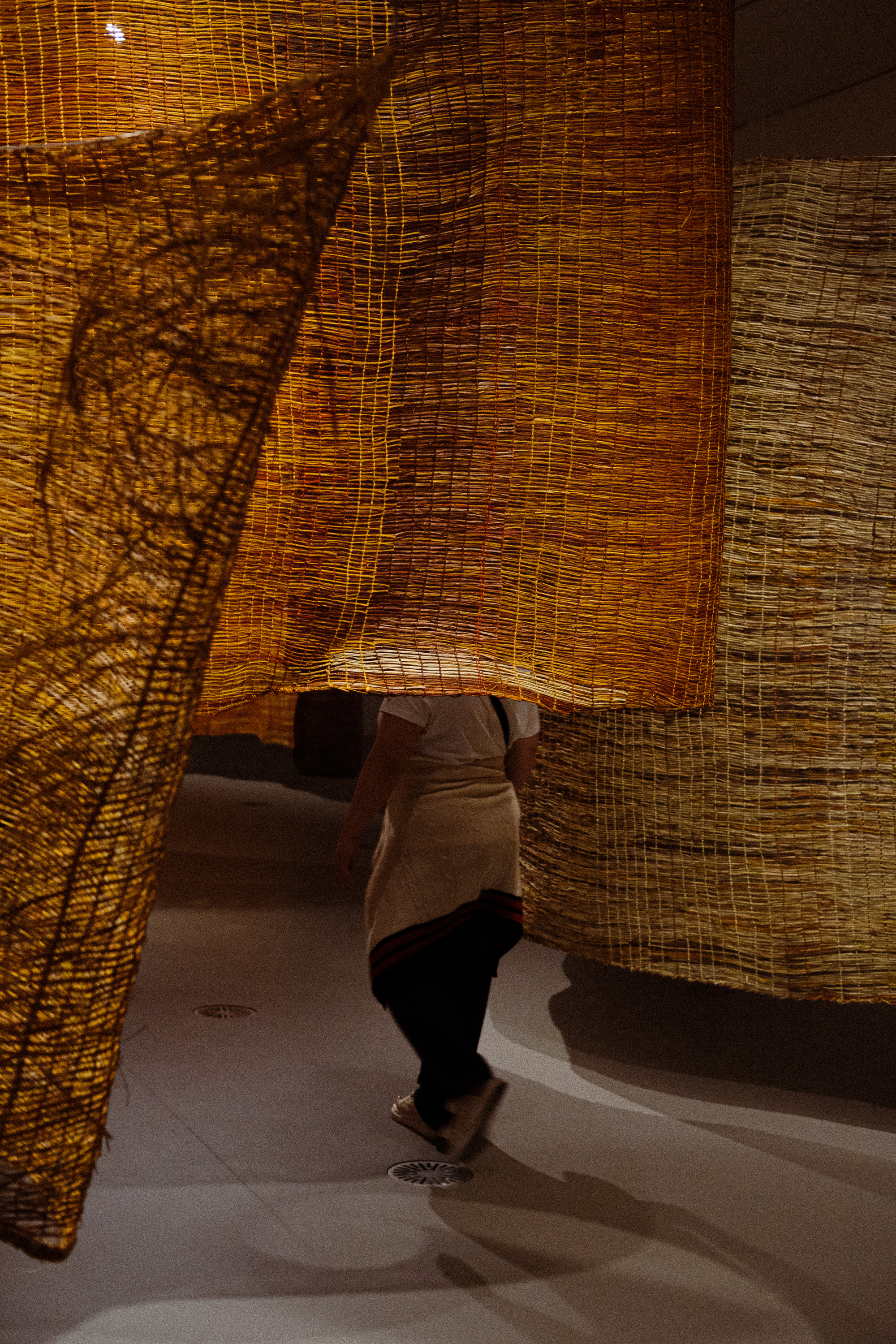 Person walking behind large woven textured yellow and orange hanging fabric panels indoors.