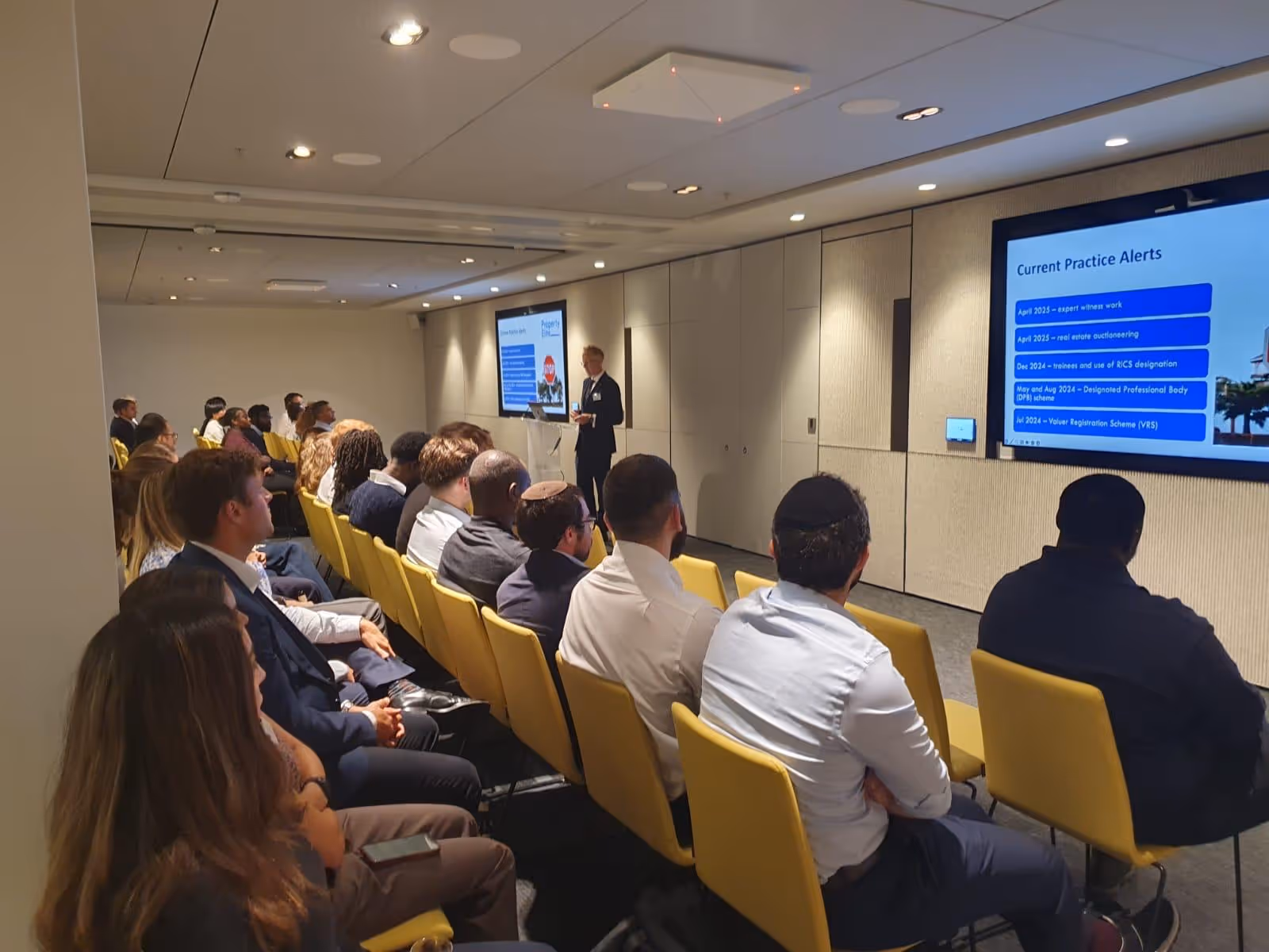 A speaker presents information on a screen to an audience seated in a conference room.
