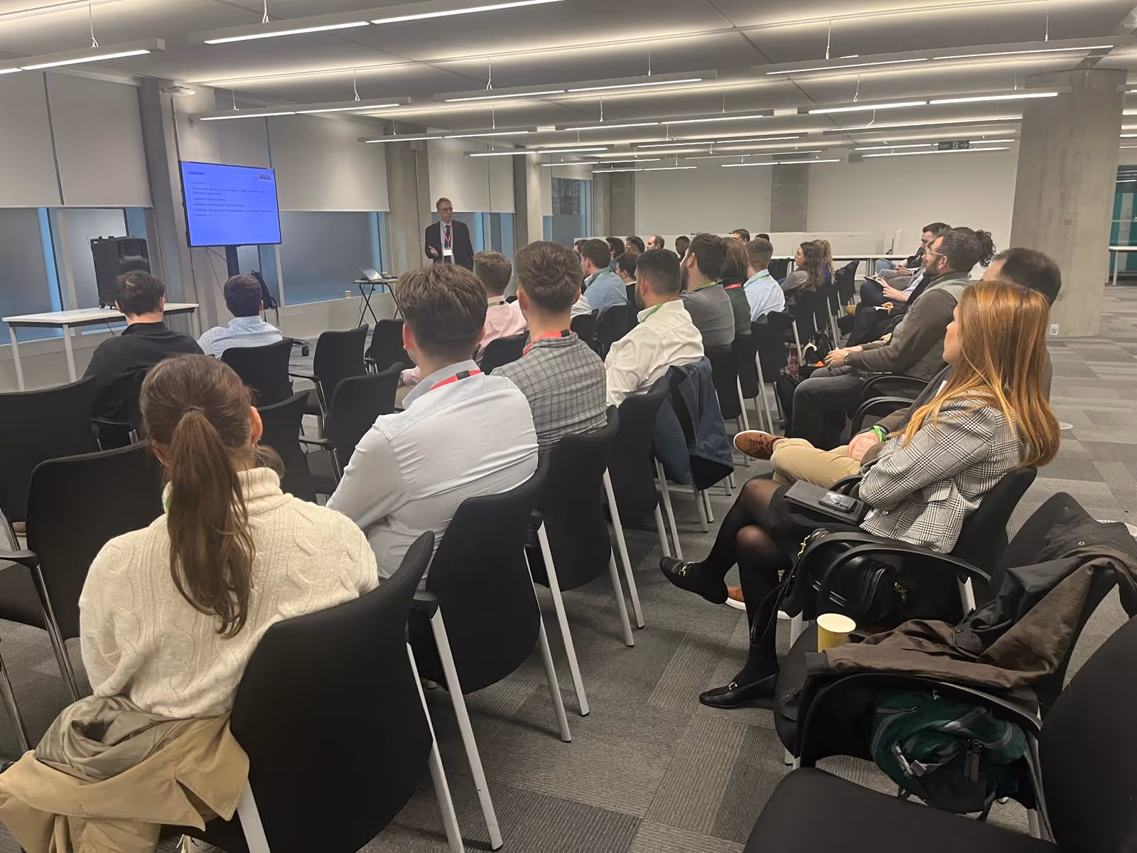 A speaker presents to an audience seated in rows in a modern, well-lit conference room.