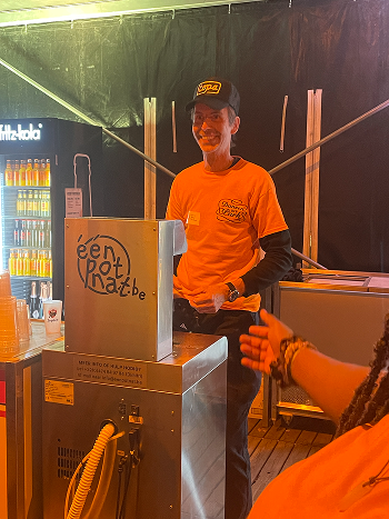 Man in orange shirt and black cap smiling behind a beer tap dispensing 'één pot nat' at an indoor event.