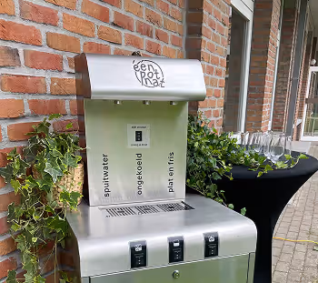 Silver beverage disposal unit with separate compartments for spuitwater, ongekoeld, and plat en fris, positioned outdoors near a brick wall and leafy green plants.