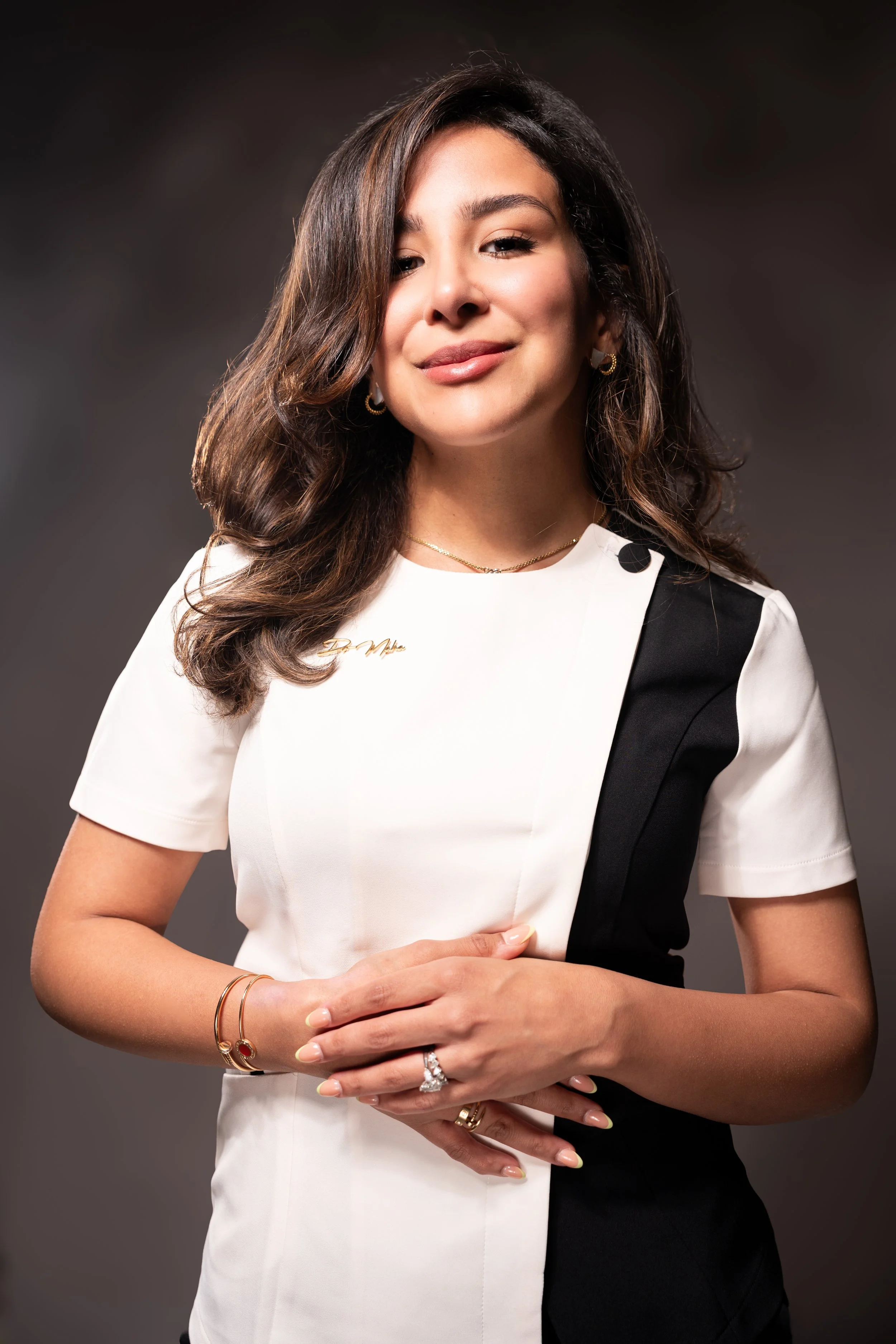 Smiling woman with wavy brown hair wearing a black and white dress and gold jewelry, posing with hands clasped.