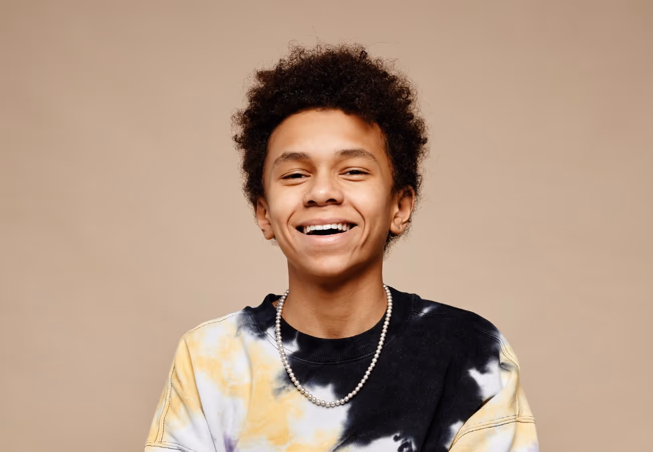 Smiling young person with curly hair wearing a yellow and black tie-dye shirt and a pearl necklace against a beige background.