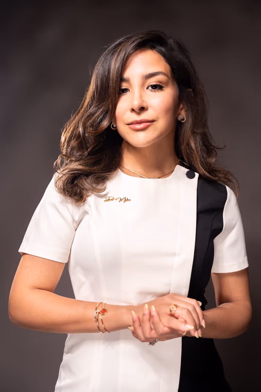 Confident woman with wavy brown hair wearing a white and black top, gold jewelry, and clasping her hands.