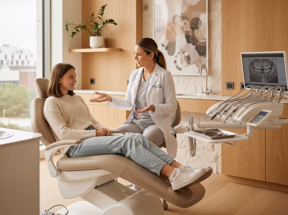 Dentist in a white coat discussing treatment with a female patient sitting in a dental chair in a modern clinic.