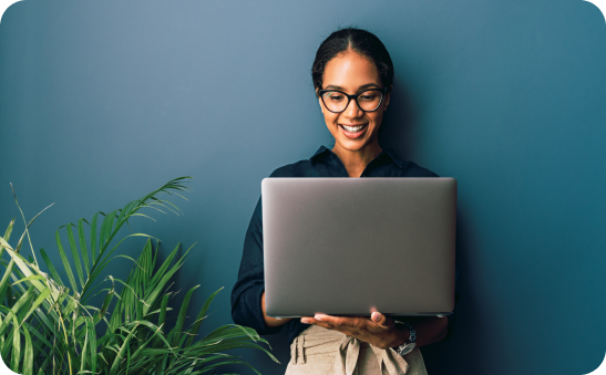 Smiling woman with glasses holding an open laptop in front of a dark blue wall with green plants nearby.