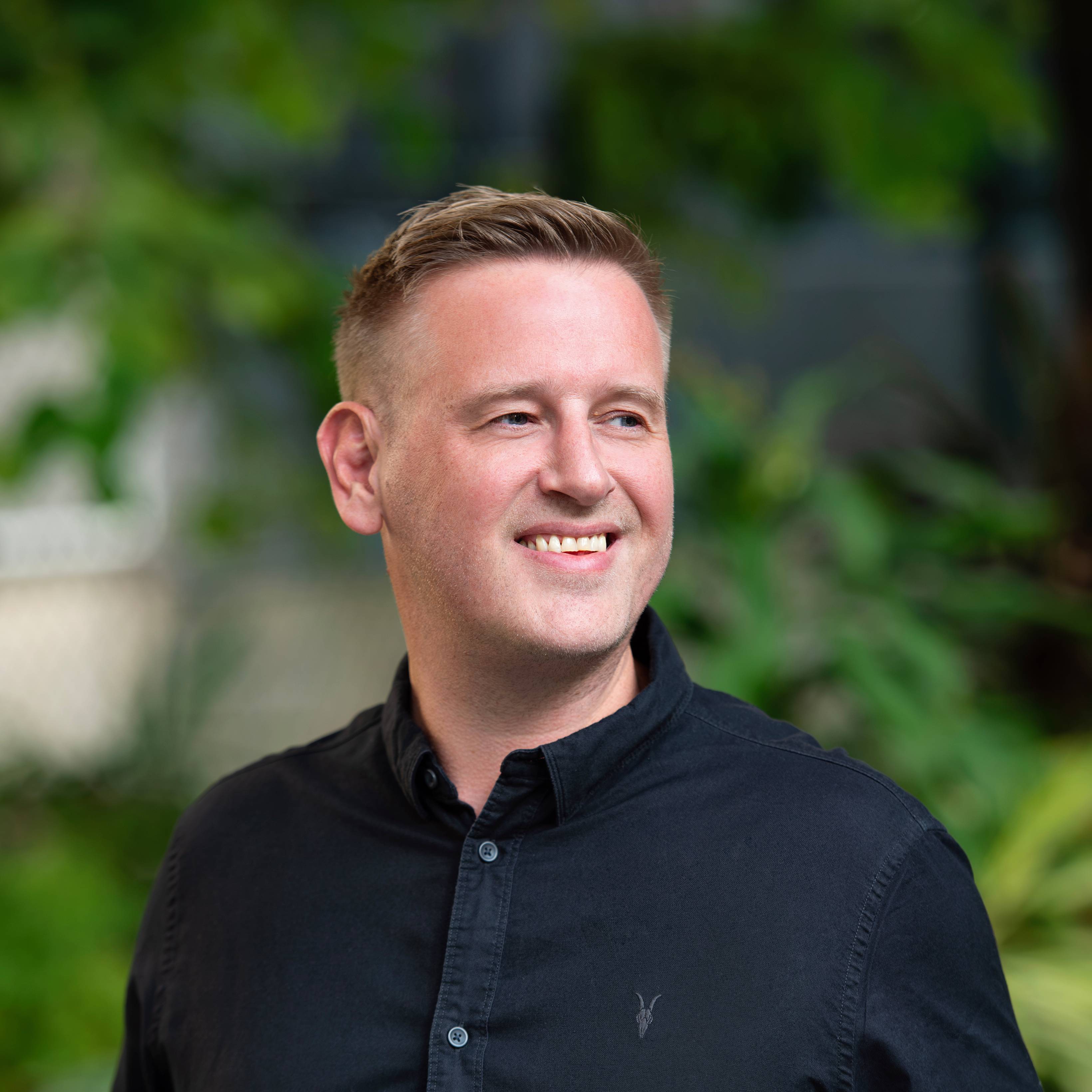 Craig Stevenson, a smiling man with short blonde hair wearing a black button-up shirt standing outdoors with green foliage in the background.