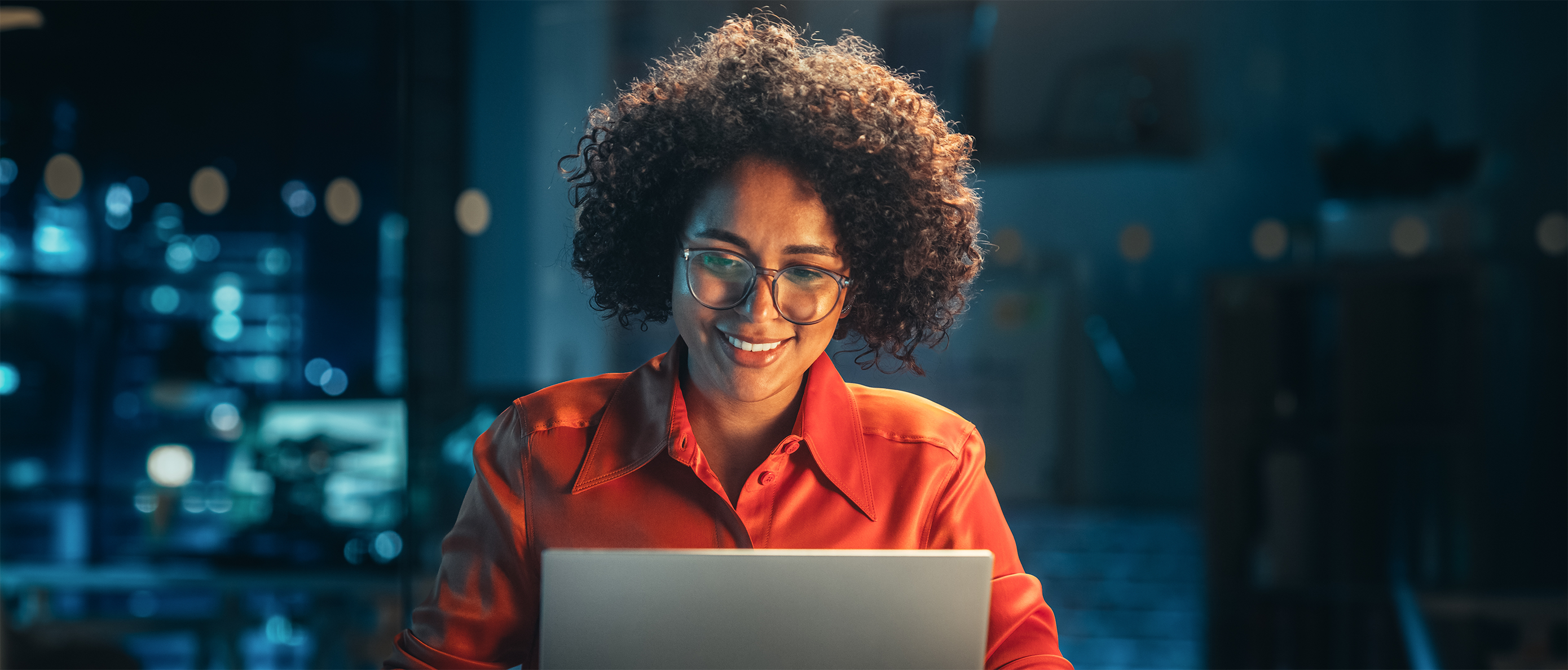 Smiling woman with curly hair and glasses working on a laptop in a dimly lit office at night.