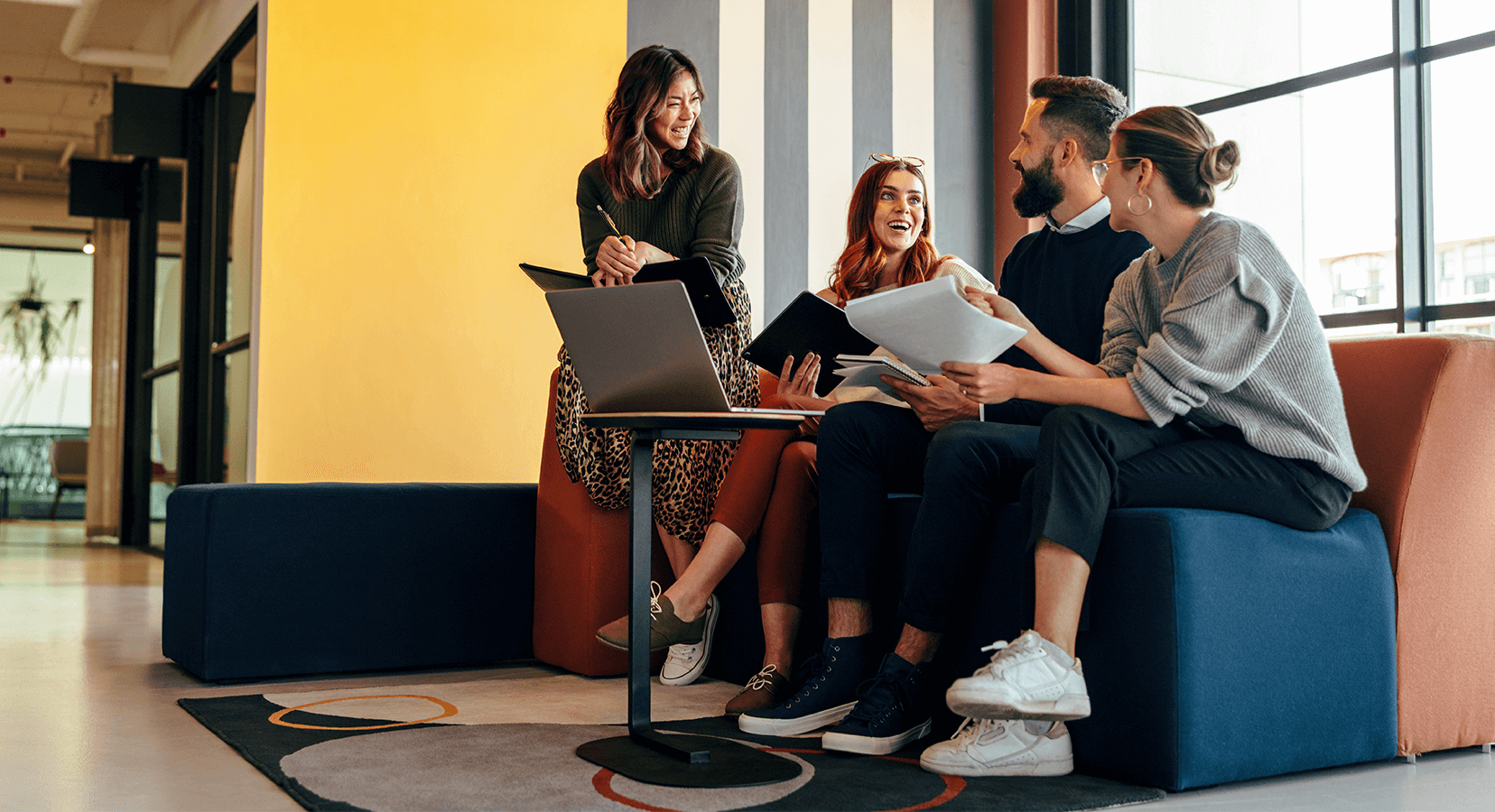 Four young professionals sitting and standing around a small table with a laptop, sharing papers and smiling in a modern office lounge.