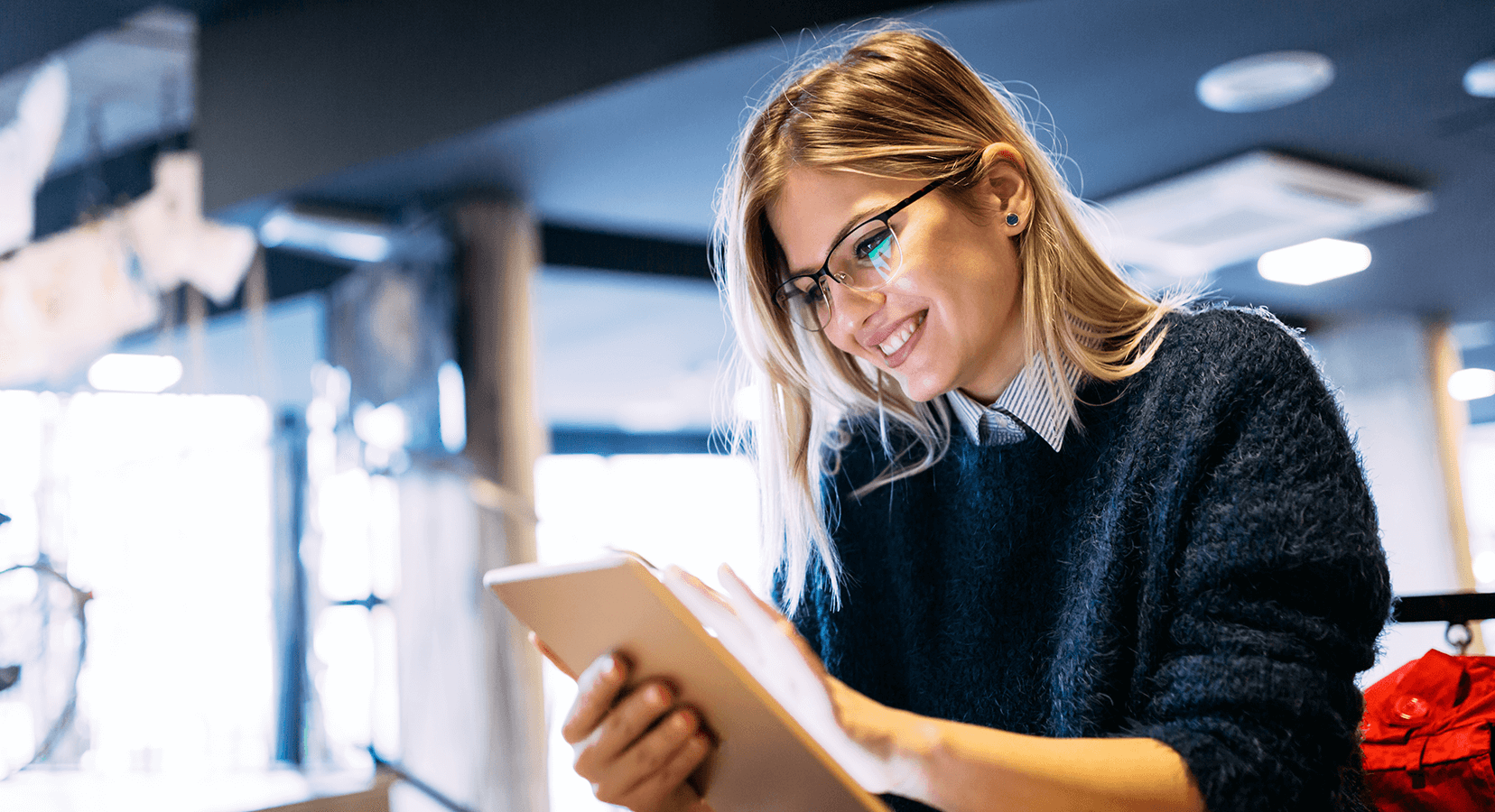 Smiling woman with glasses using a tablet in a modern, bright indoor setting.