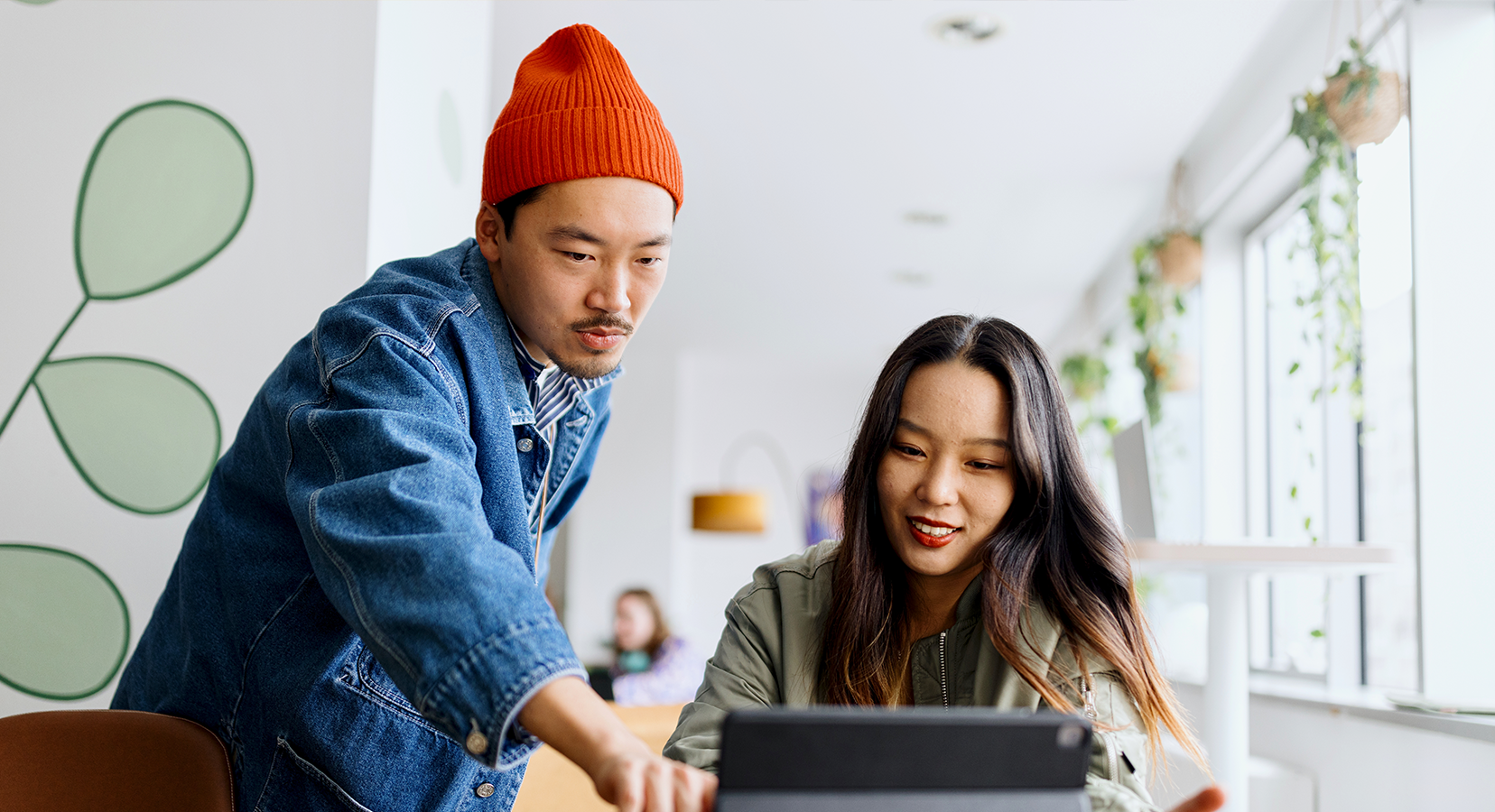 A man in a red beanie and denim jacket points at a tablet while a woman in a green jacket smiles and looks at the screen in a bright, modern room.