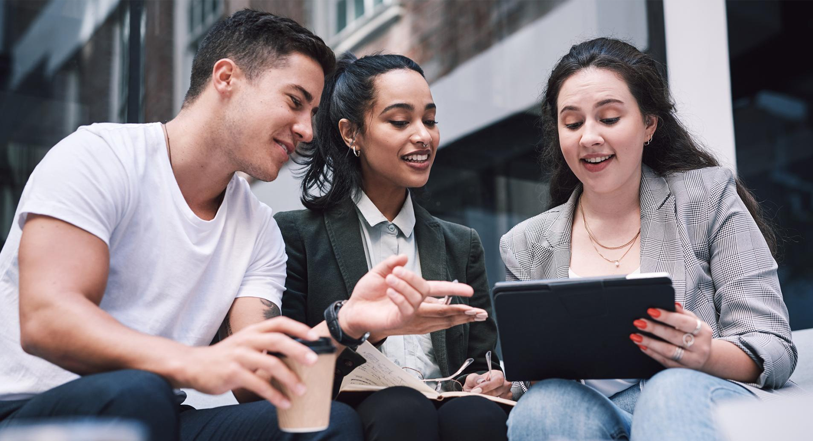 Three young professionals sitting outdoors and smiling while looking at a tablet during a discussion.