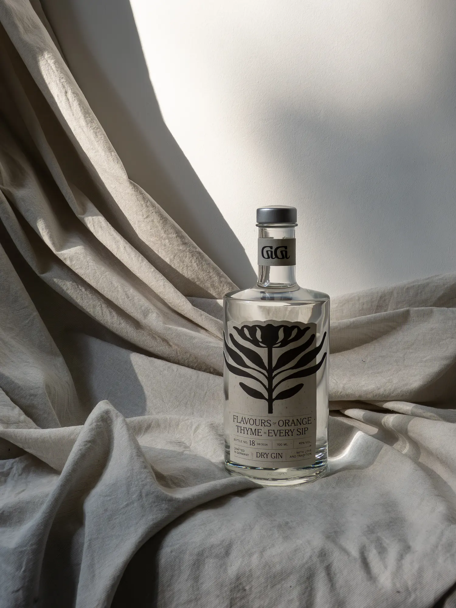 Klare Flasche mit trockenem Gin mit schwarzem Blumenmuster und Text über Orangen- und Thymianaromen, auf beigem Stoff mit Sonnenlicht und Schatten platziert.