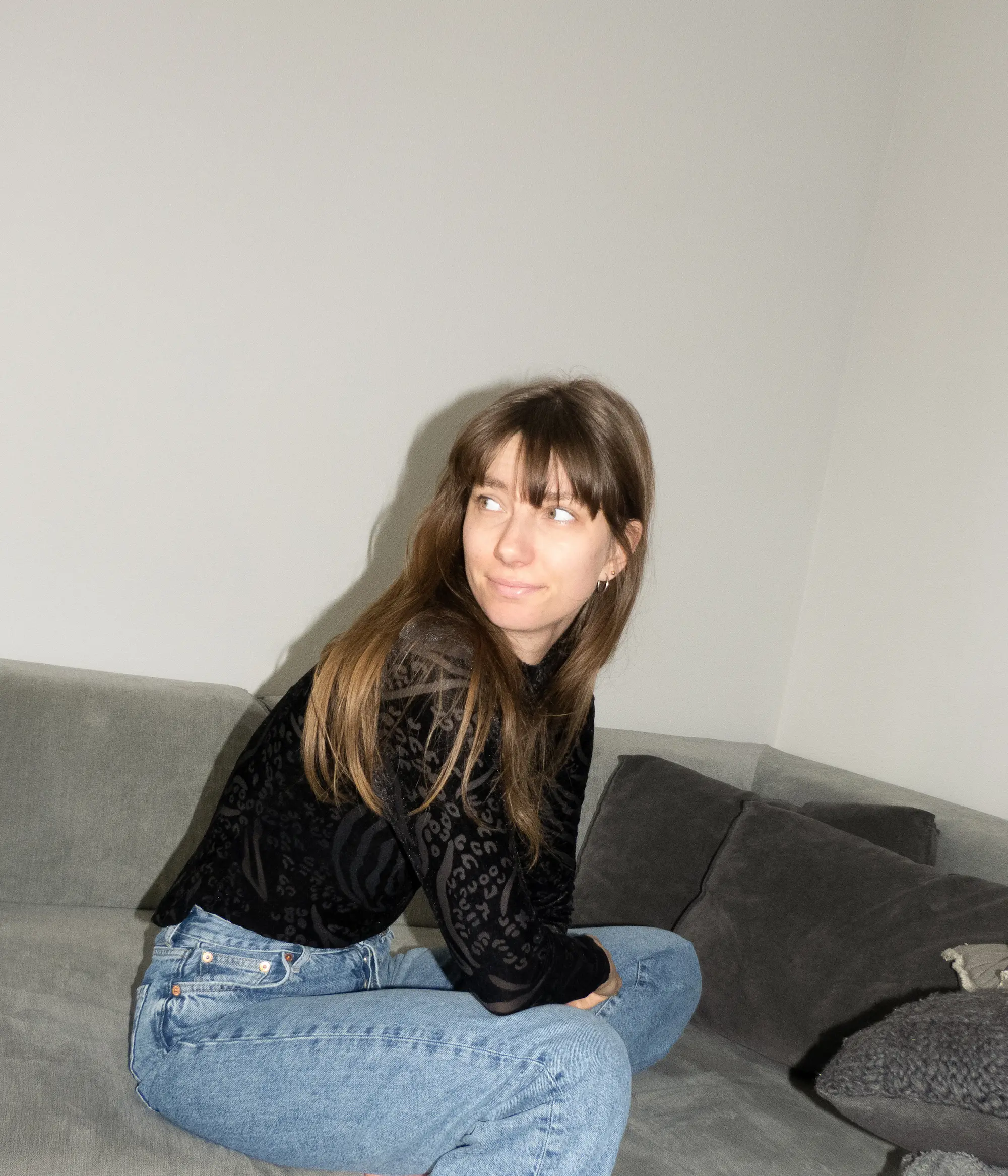 Junge Frau mit langen braunen Haaren, die mit gekreuzten Beinen auf einem grauen Sofa sitzt und nach links schaut.