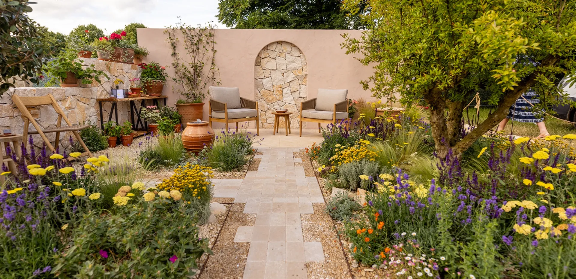 Pathway through a colorful garden with yellow and purple flowers leading to two cushioned wooden chairs and a small table against a stone and plaster wall.