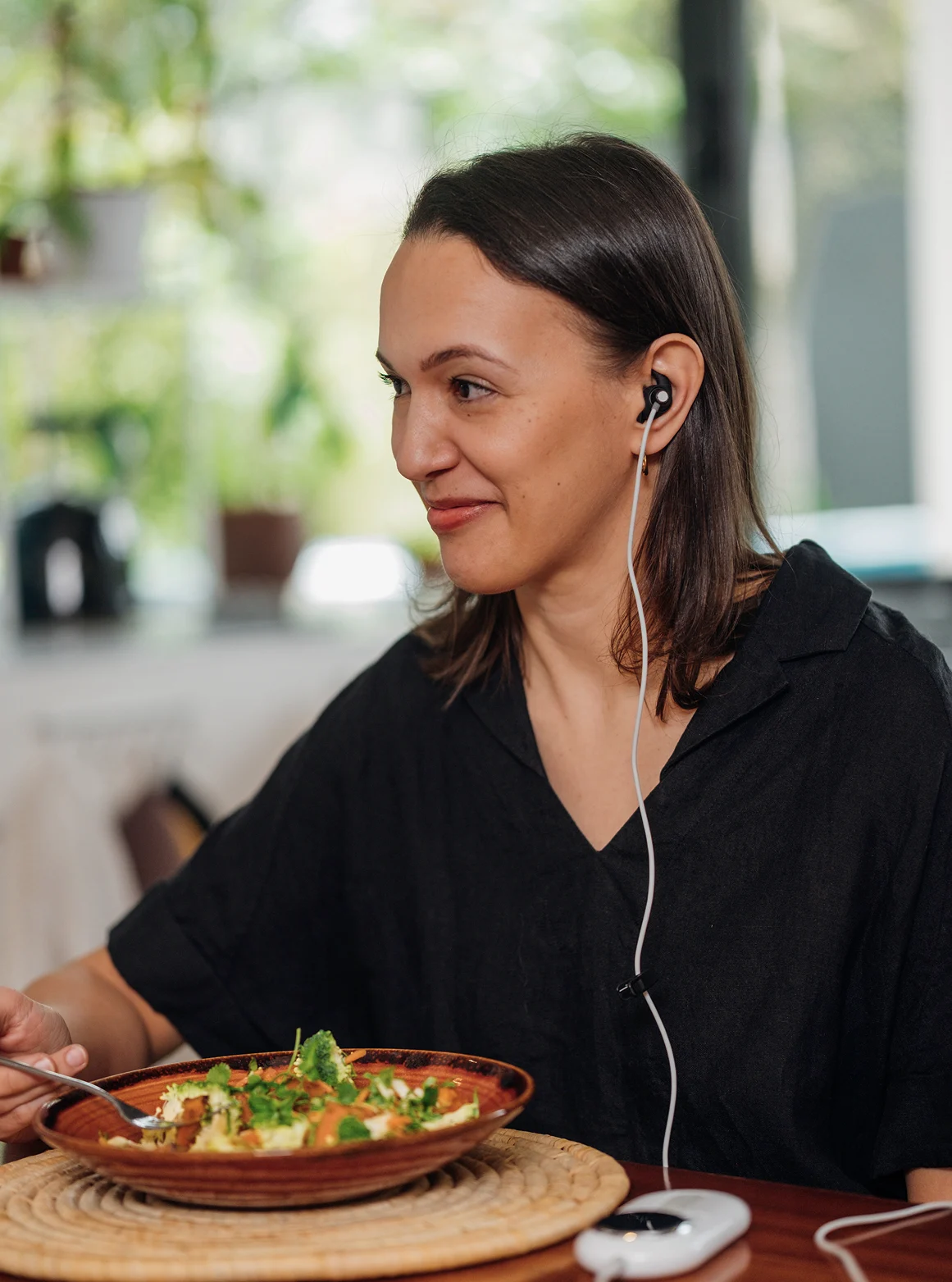 Woman wearing black shirt and black earphones, smiling while holding a fork over a plate of salad with broccoli, sitting at a table.