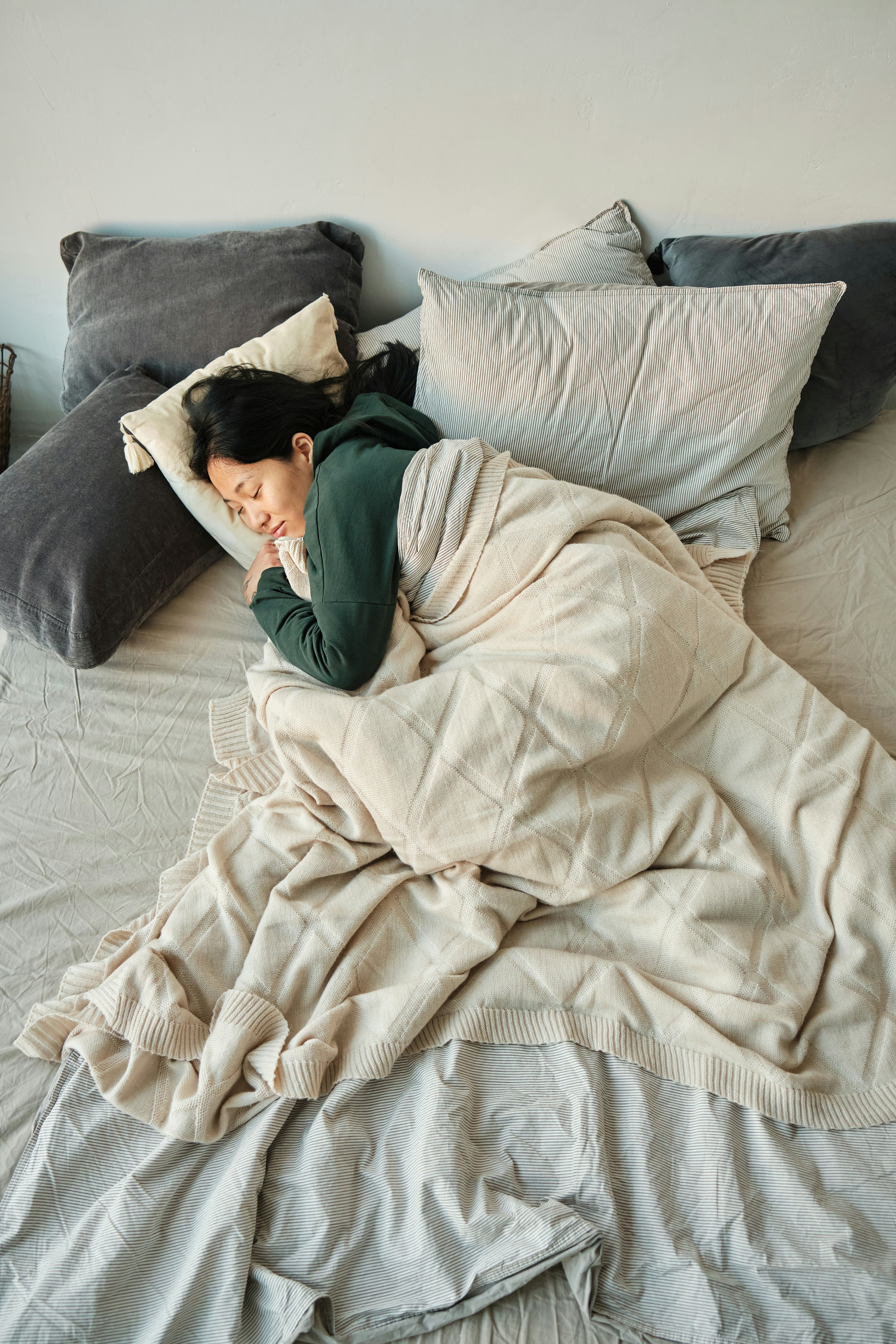 Woman sleeping on a bed wrapped in a beige blanket with multiple pillows.