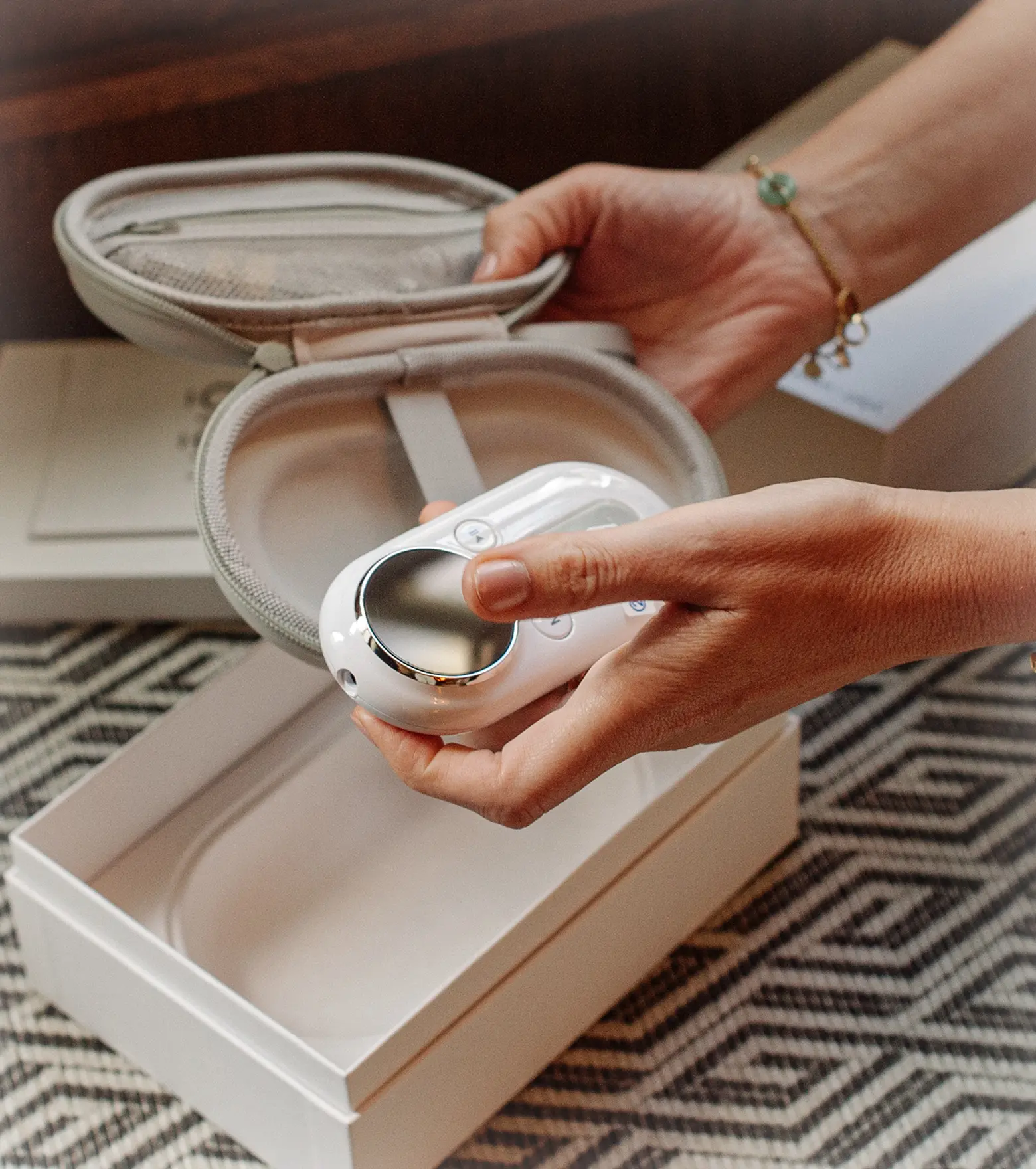 Hands holding a white electronic device with buttons and a circular metallic surface above an open protective carrying case and a white box on a patterned surface.