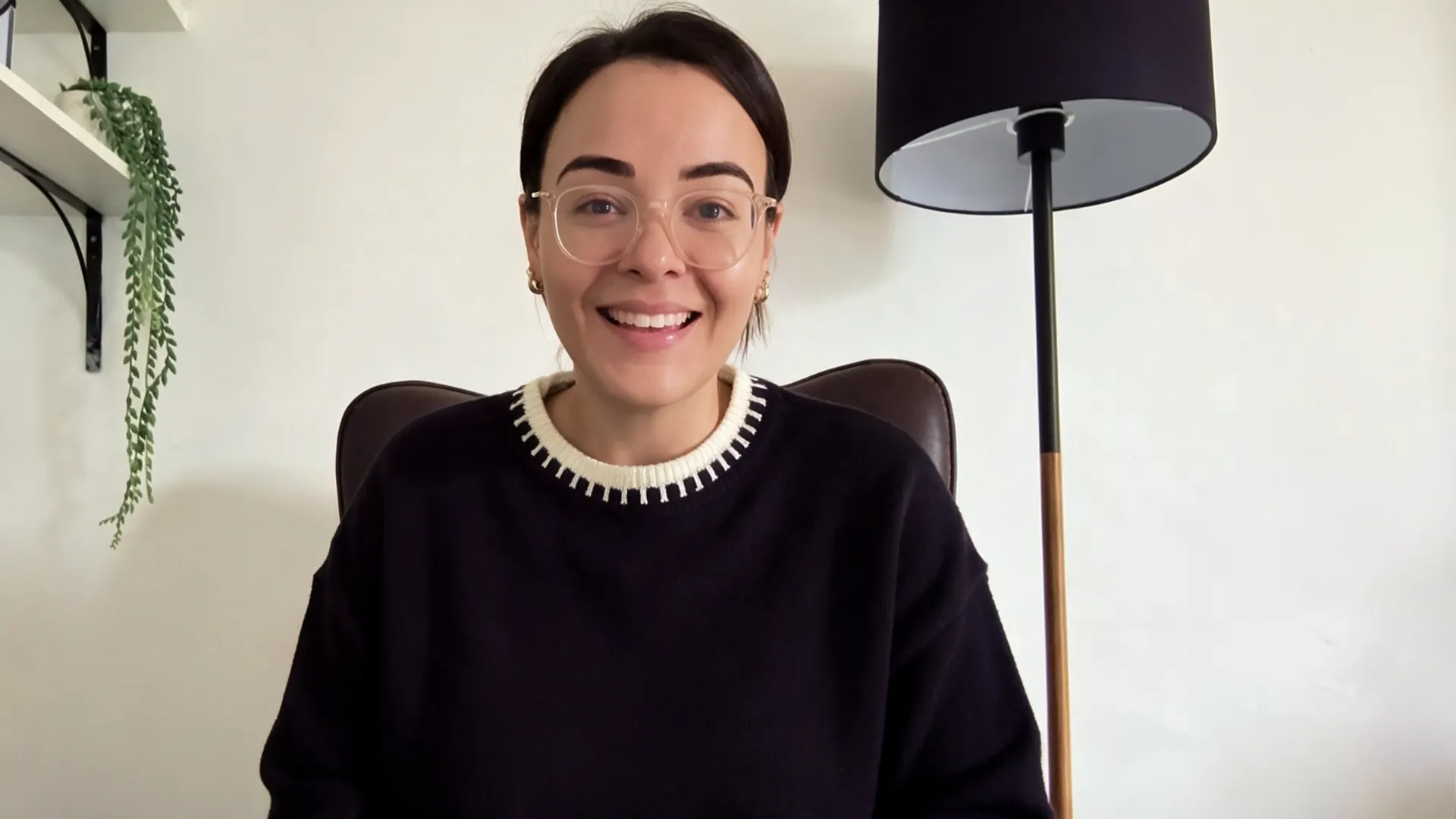 Smiling coach with short dark hair and glasses sitting in a chair in a room with a lamp and plant.