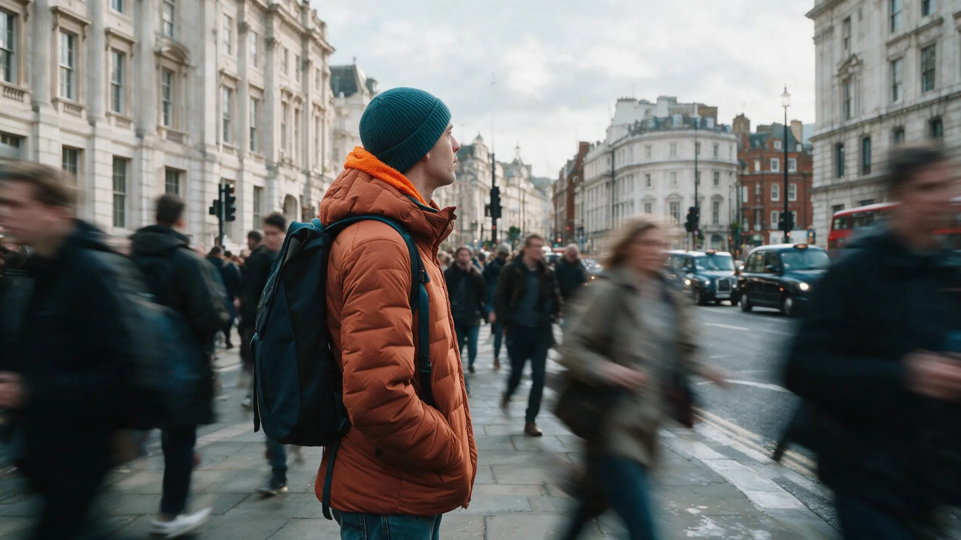 Man in orange jacket and blue hat standing on busy London sidewalk with blurred moving pedestrians.
