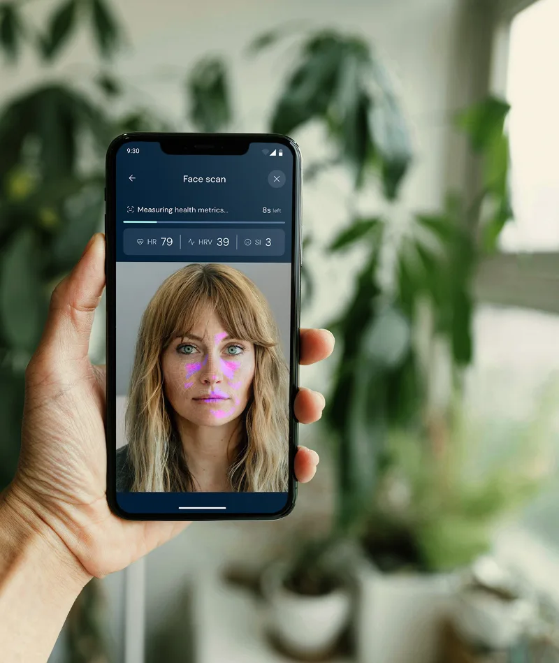 Hand holding phone showing a face scan app measuring a woman’s health metrics with facial highlights.