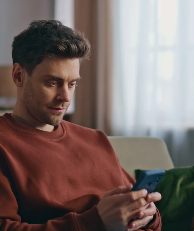 Man in rust-colored sweater chatting with his coach on his phone while sitting on a couch.