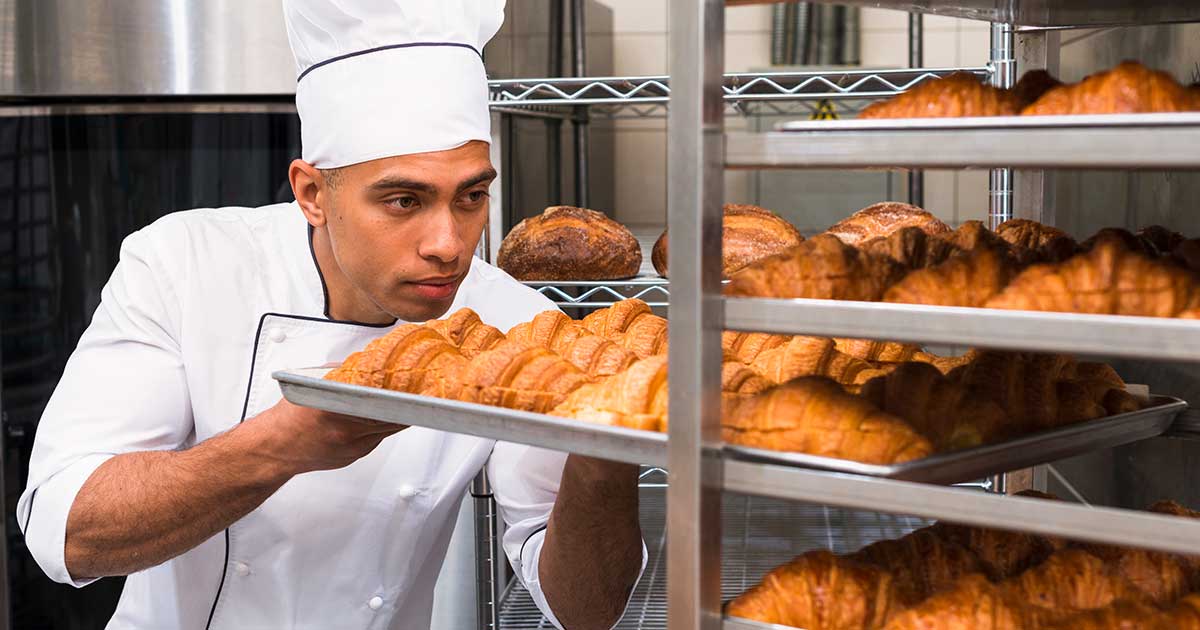 Panadero con uniforme blanco inspecciona una bandeja llena de croissants recién horneados en una cocina profesional.