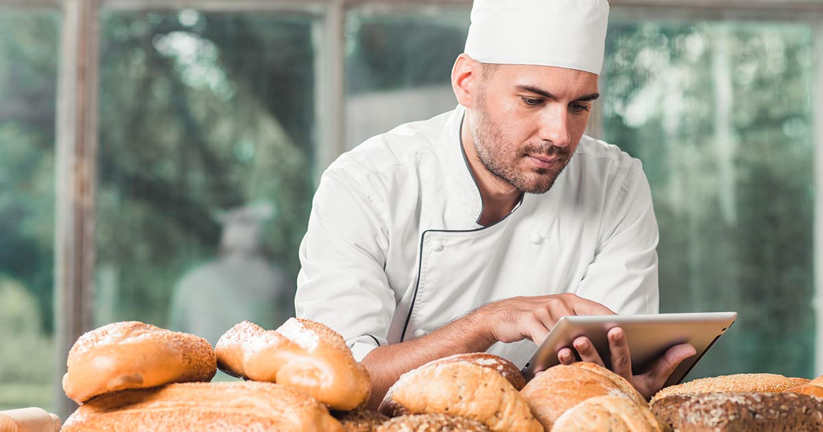 Panadero con uniforme blanco usando una tableta digital detrás de una mesa con variedad de panes.